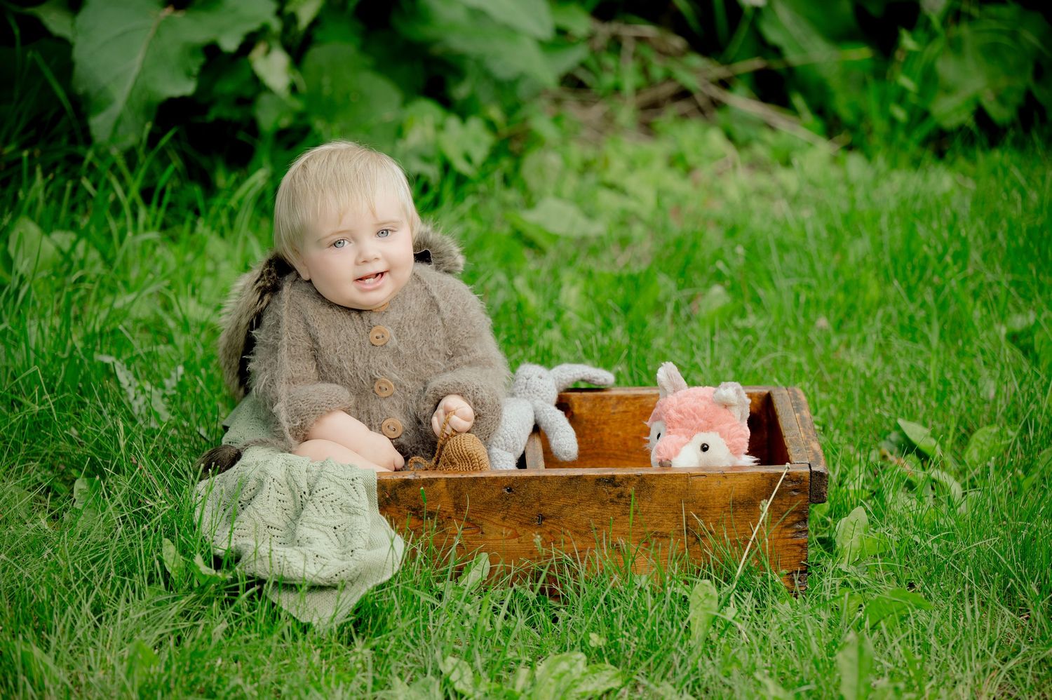 Smiling baby in brown sweater sits on grass beside wooden crate with stuffed animal toys outdoors. Milestone and Birthday Photographer in Ce