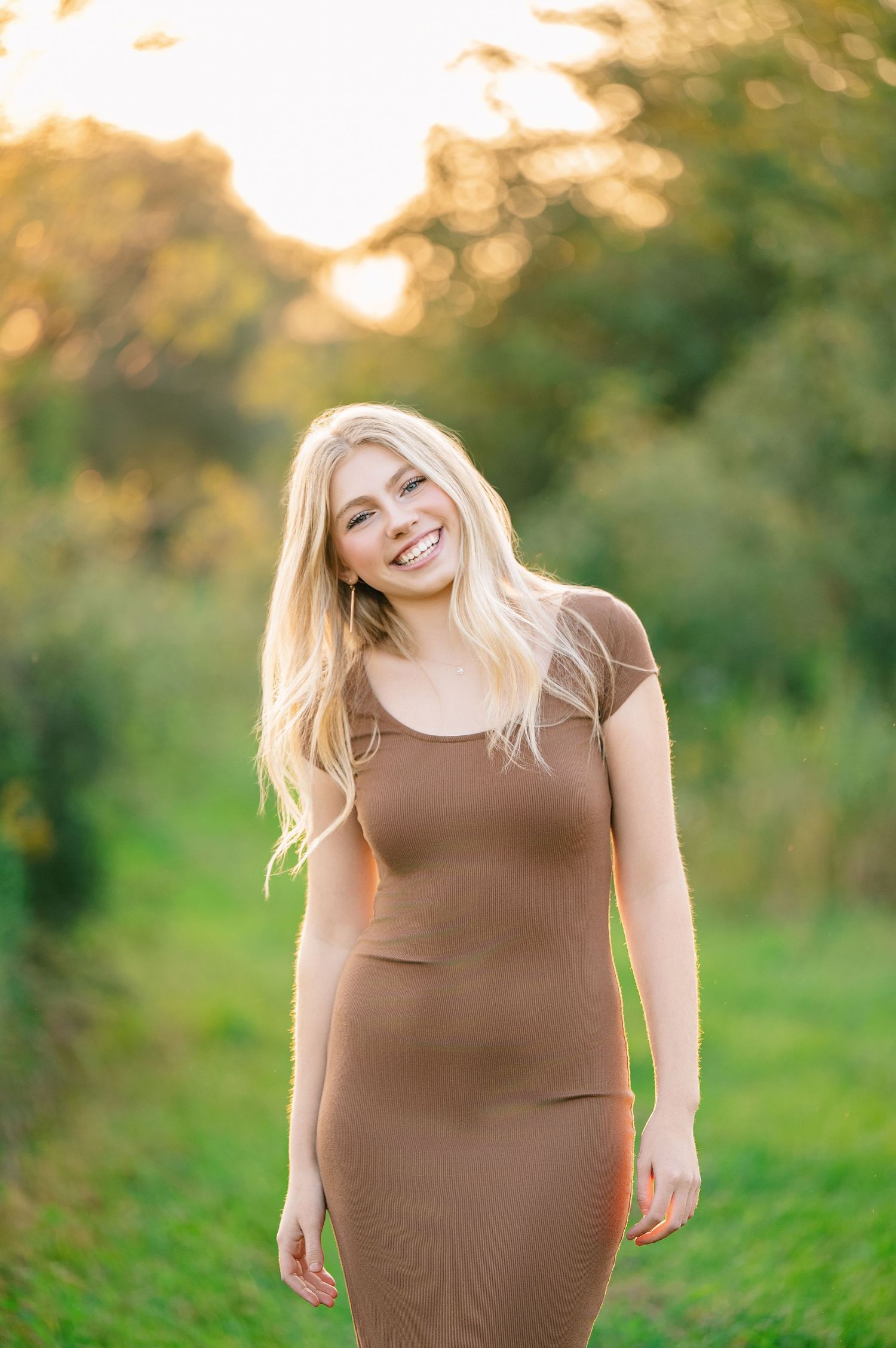 Central MN Senior Photos - Smiling blonde woman in brown ribbed dress standing outdoors in golden hour sunlight with green trees behind her.