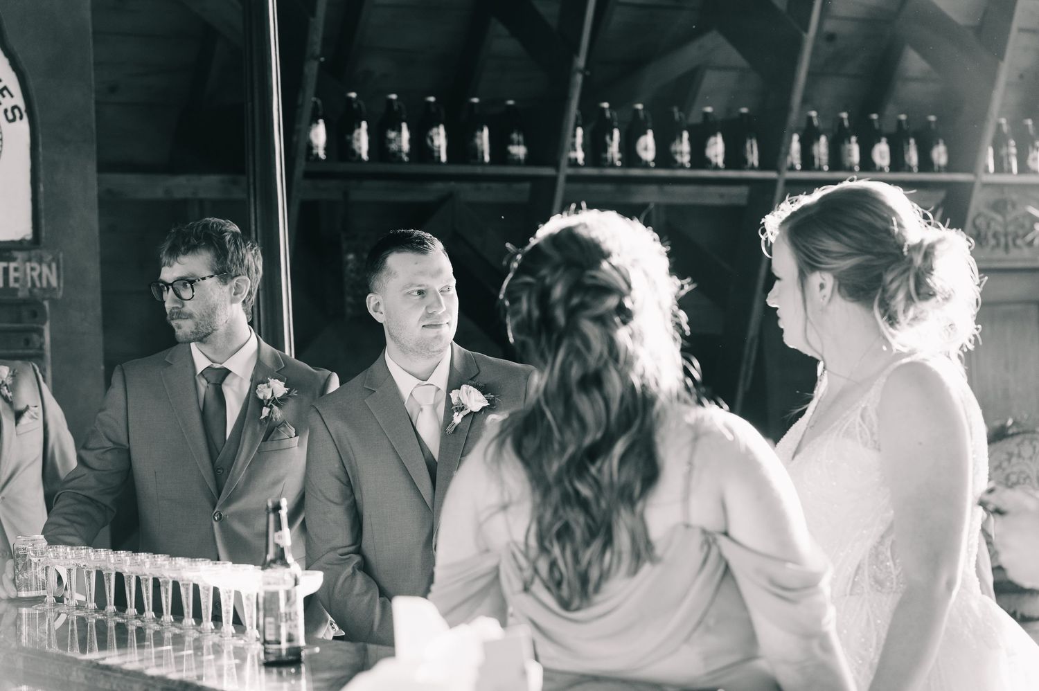 Black and white photo of groom and guests conversing at bar with bottle display backdrop.