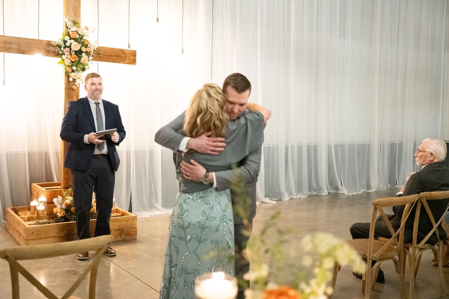 Emotional hug between guests during wedding ceremony with officiant and cross backdrop.