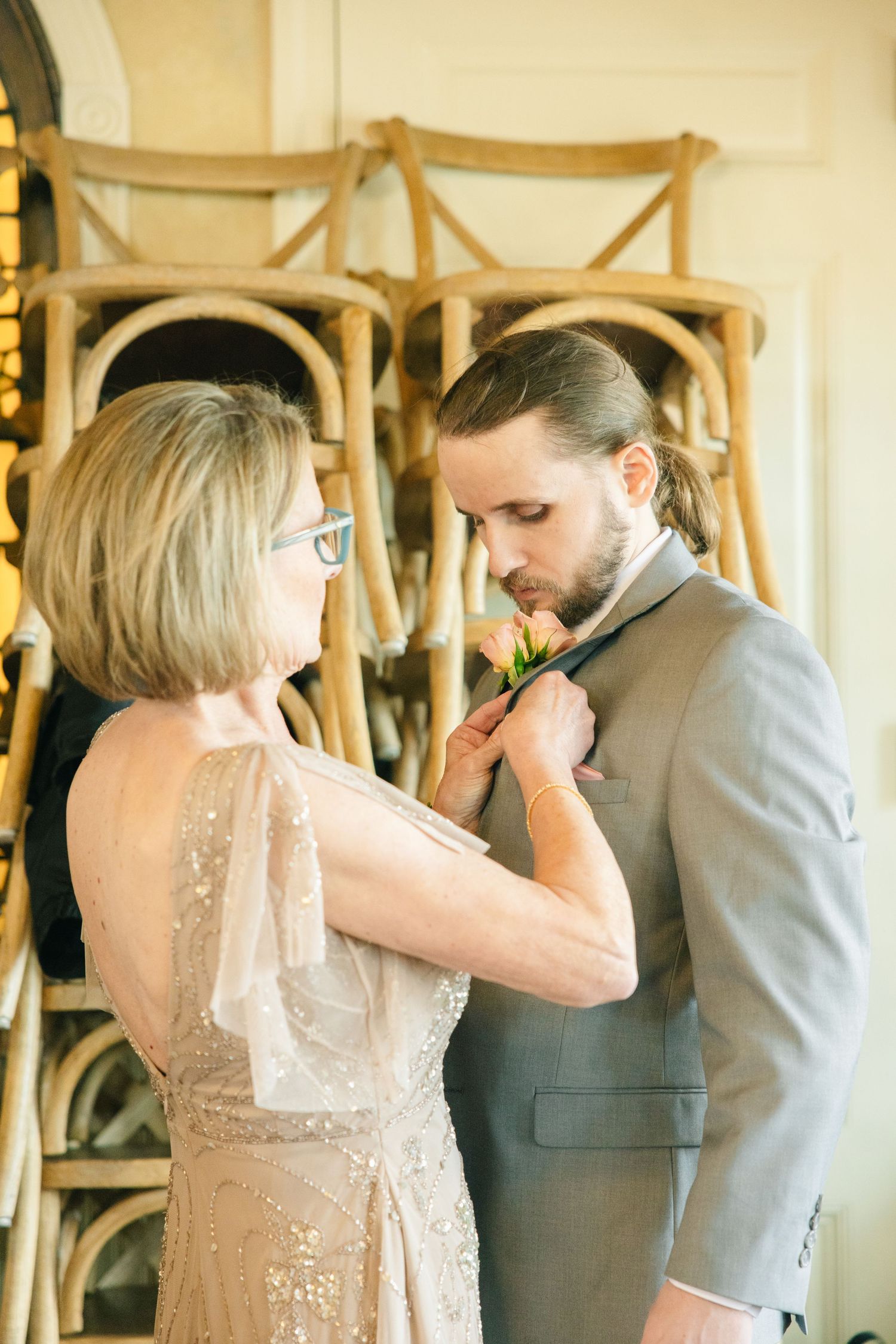 Mother pinning boutonniere on groom in gray suit before wedding ceremony.