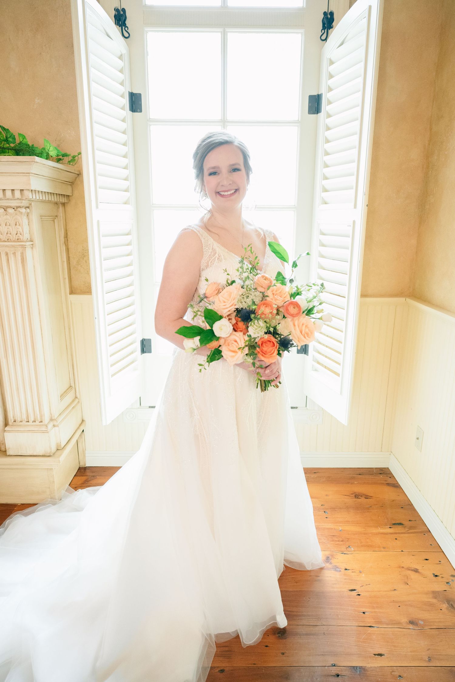 Smiling bride in white gown holding peach floral bouquet standing before shuttered window on hardwood floor.