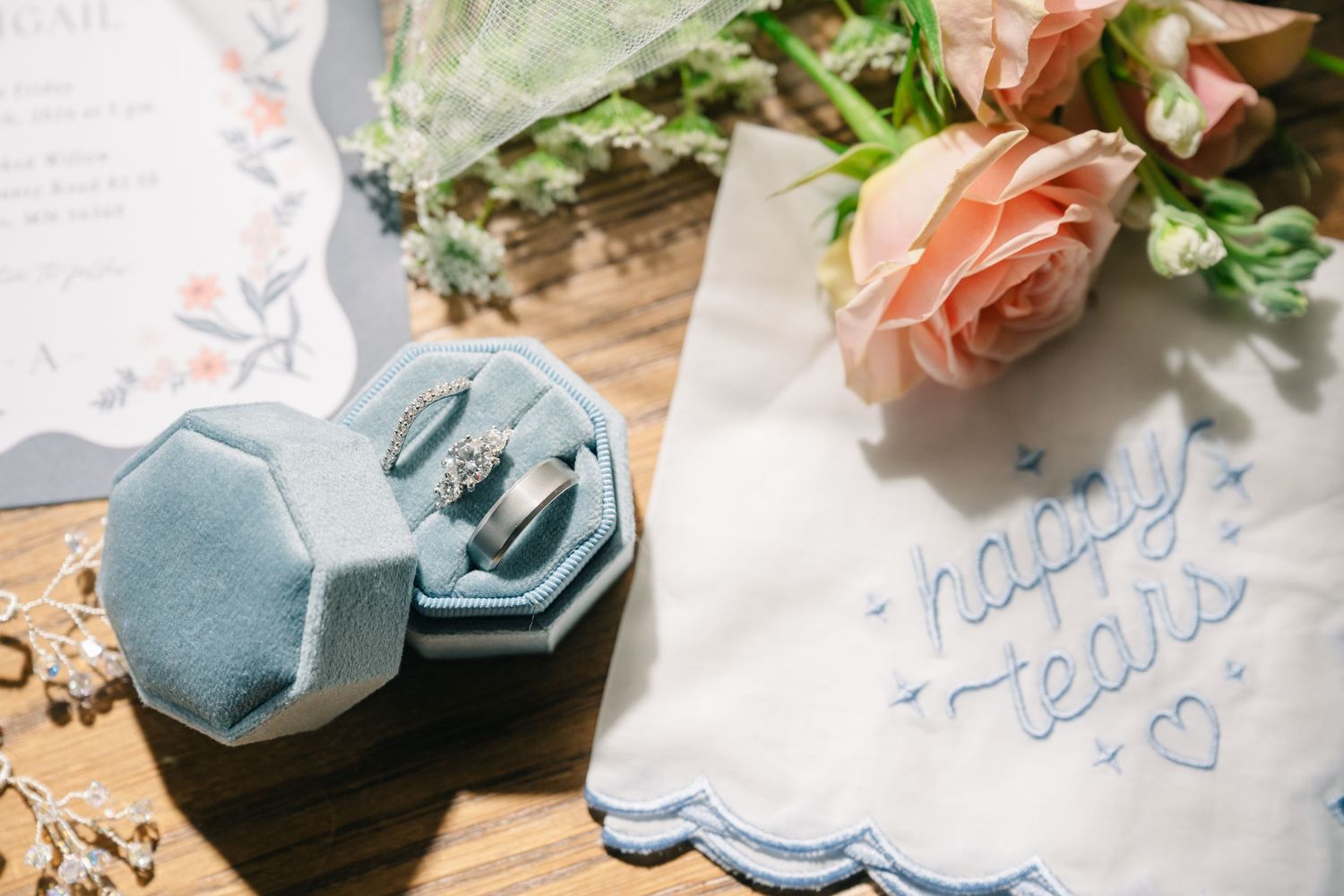 crooked willow weddiBlue velvet ring box with diamond rings beside pink roses, wedding invitation, and happy tears embroidered handkerchief.