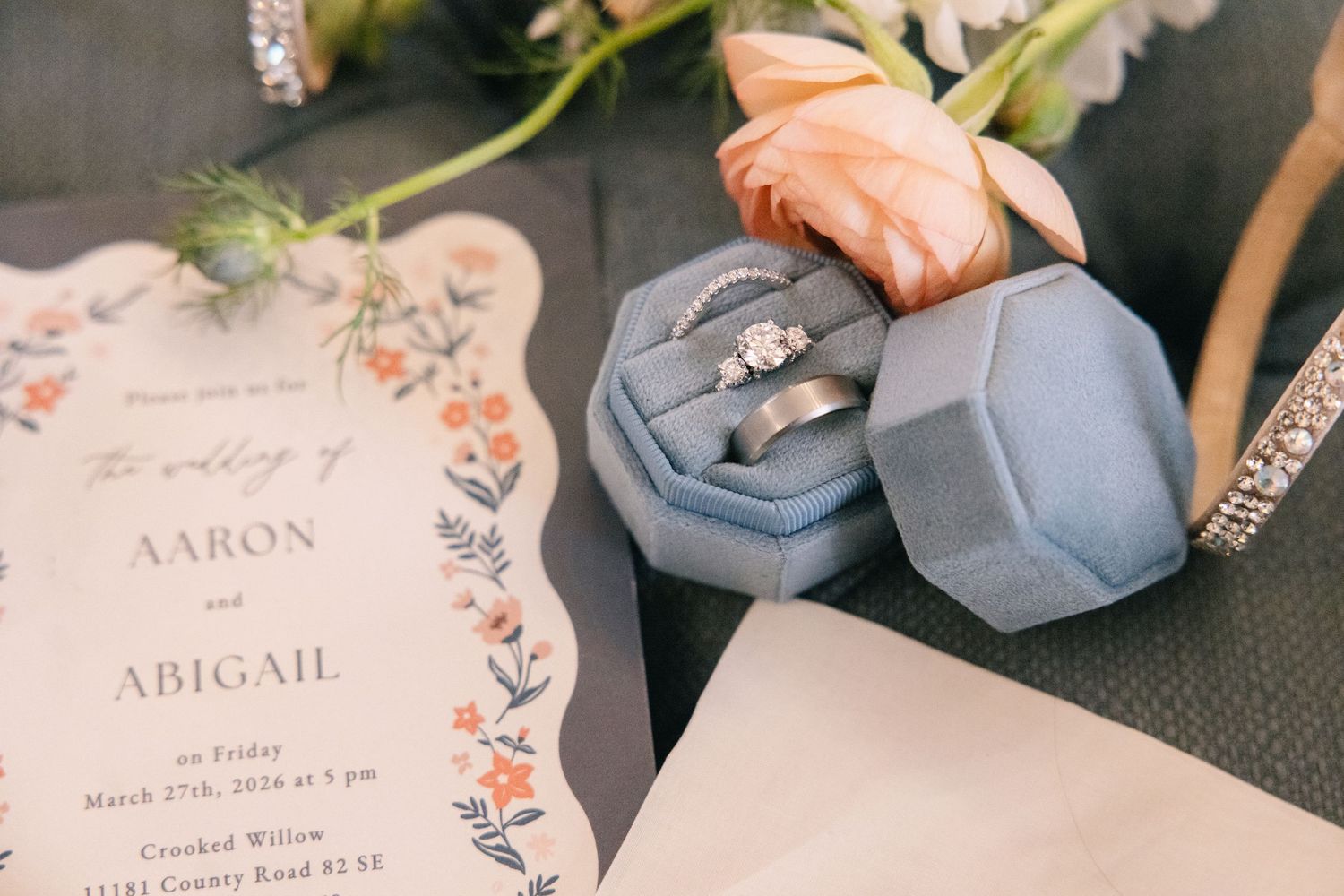 crooked willow weddings Wedding rings in a blue velvet box beside a floral invitation for Aaron and Abigail's wedding with a peach rose.