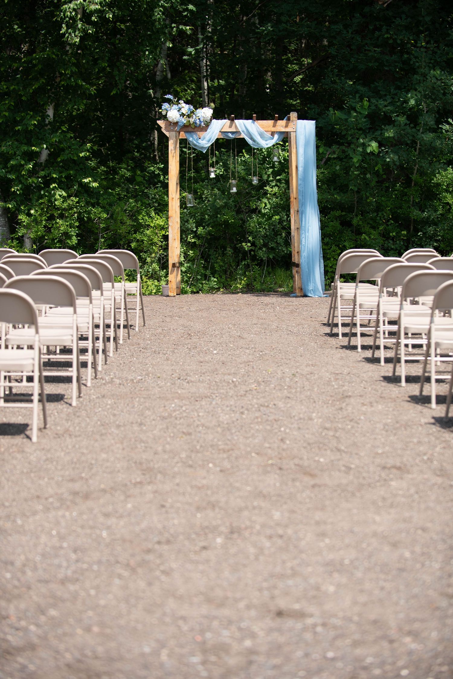 Outdoor wedding ceremony setup with wooden arch decorated in blue and white fabric and rows of white chairs on gravel path. Zimmerman MN Wed