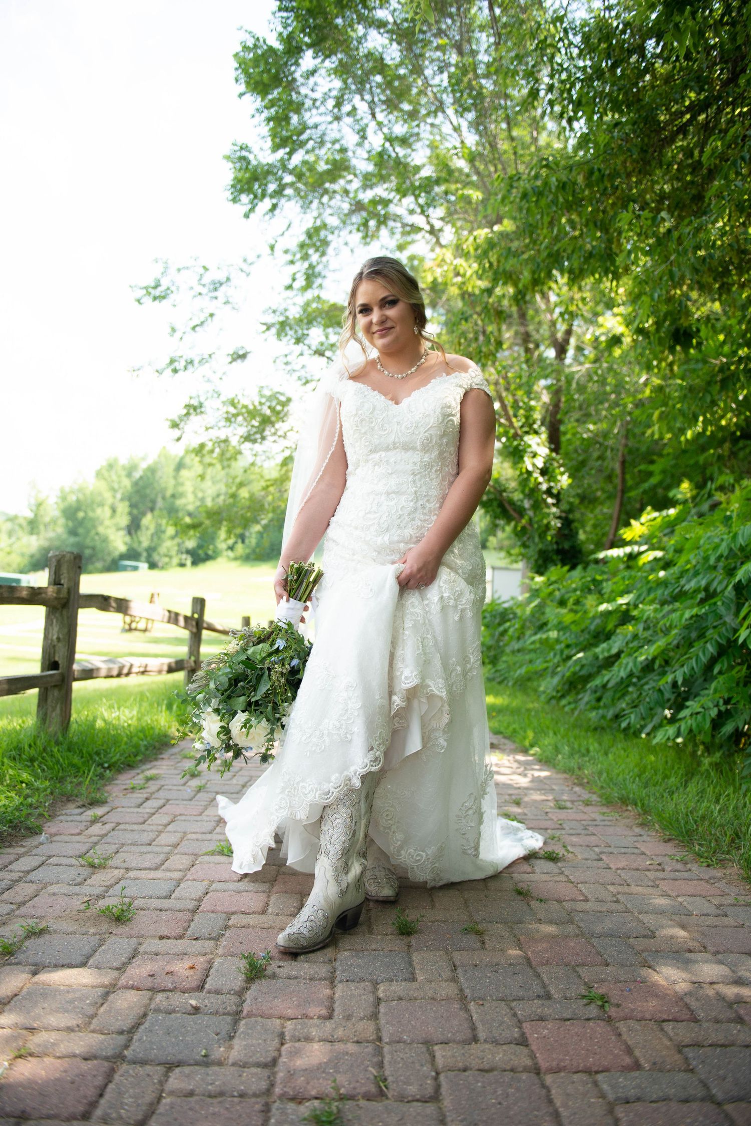 Bride in lace gown poses on rustic brick pathway surrounded by greenery. Zimmerman MN Weddings