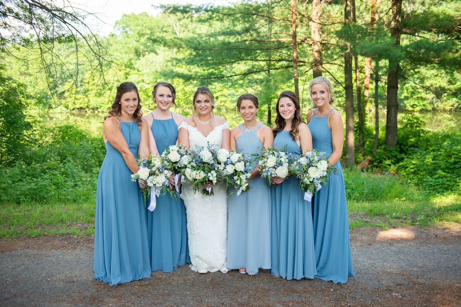 Row of bridesmaids in blue dresses holding white floral bouquets in a forest setting. Zimmerman MN Weddings