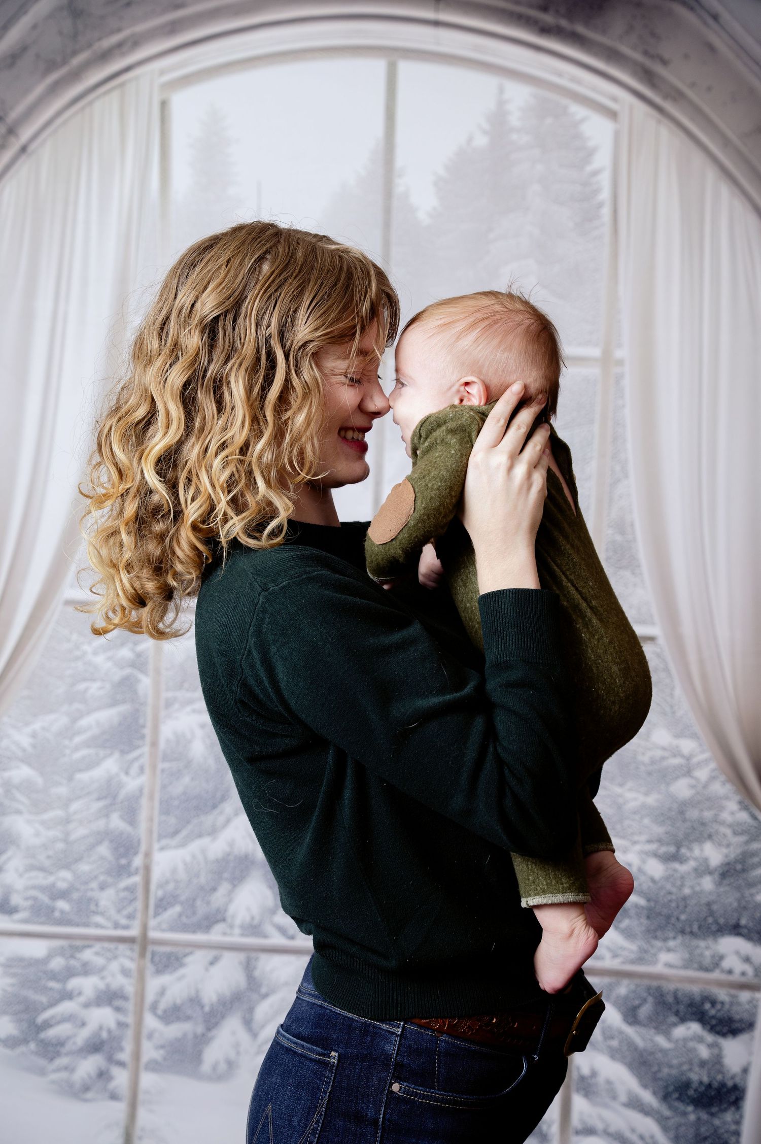 A tender moment by a snowy window as a woman in a black sweater holds and gently kisses her infant wearing green. Princeton MN Milestone, Ma