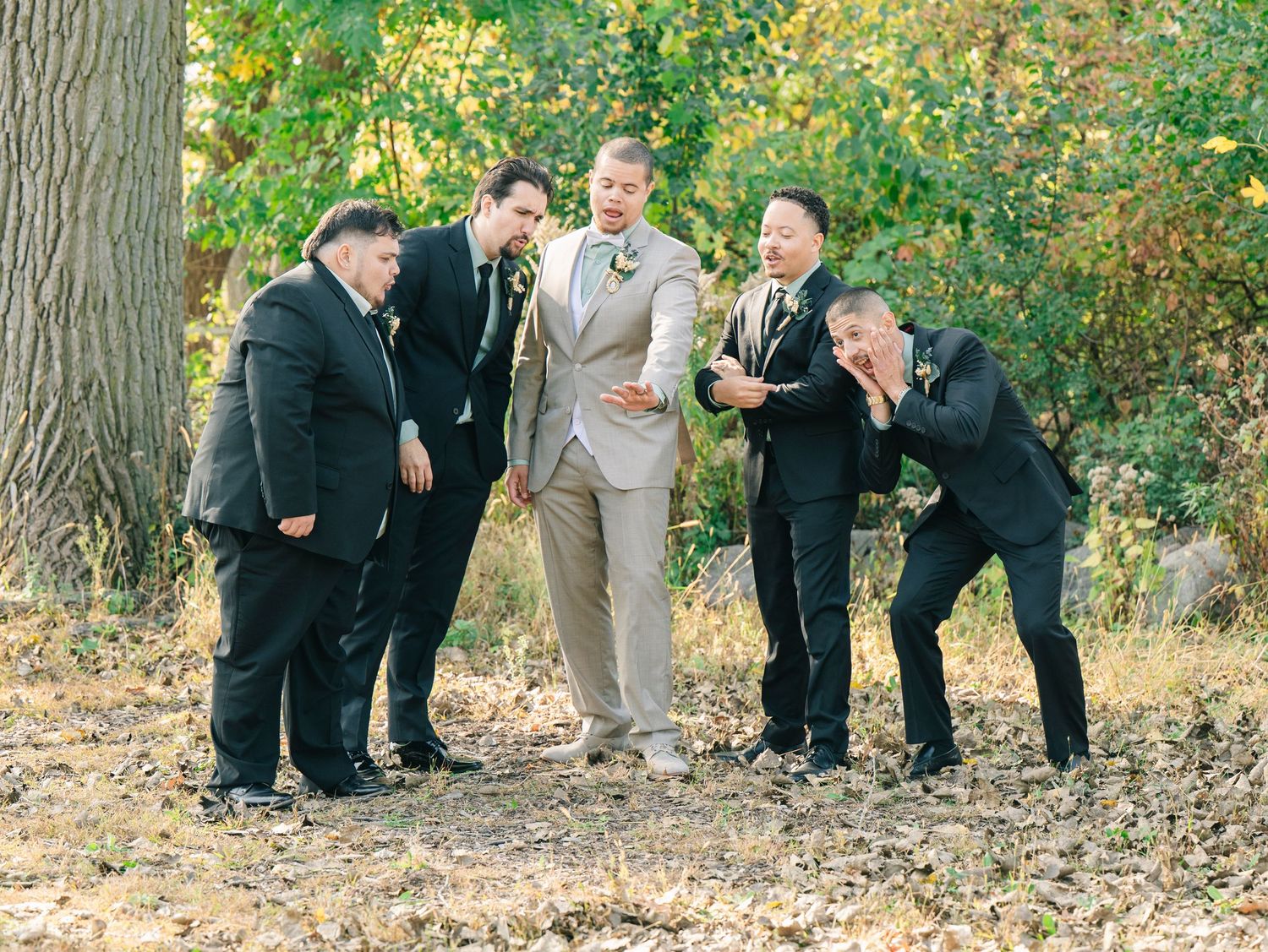 Groom in tan suit poses with groomsmen in black suits outdoors among autumn trees for a wedding photo.