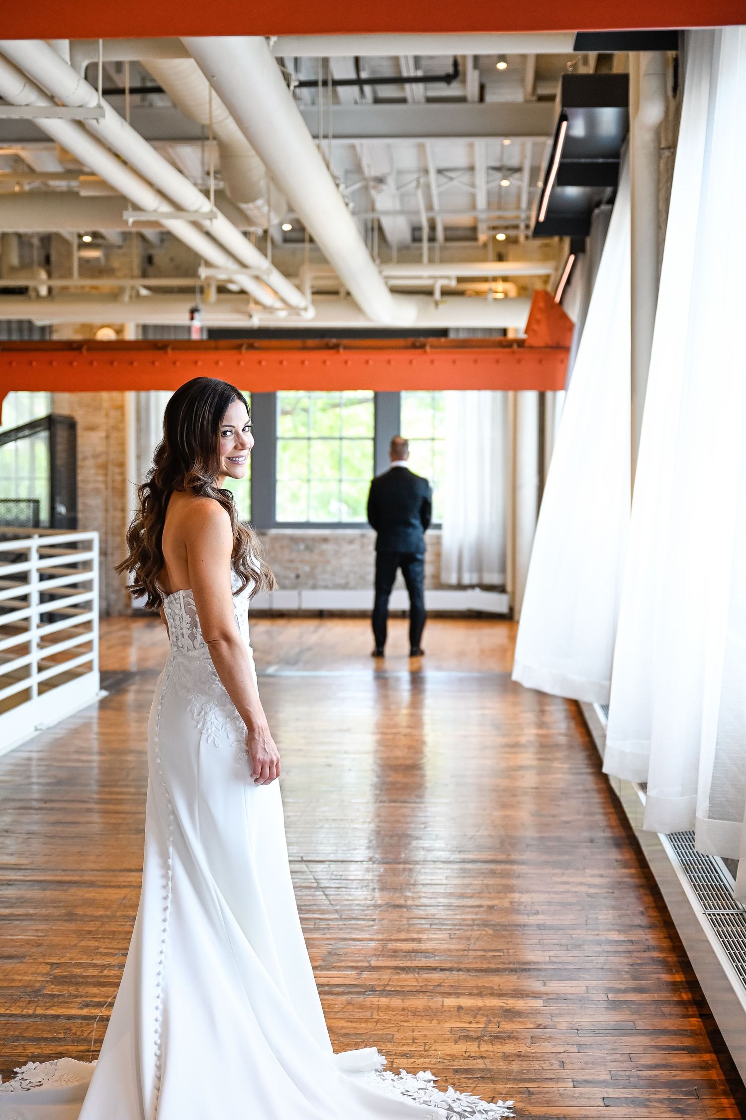A bride in a flowing white gown with long train stands in a bright industrial-style room with wooden floors.