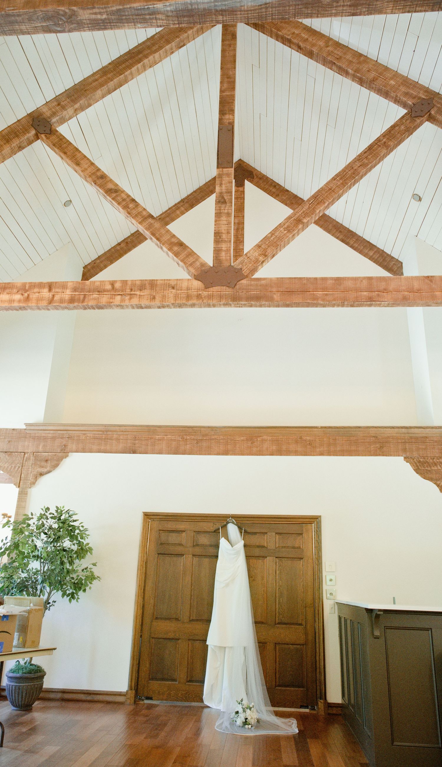 Wedding dress hanging over wooden doors beneath vaulted ceiling with exposed timber beams in elegant venue.