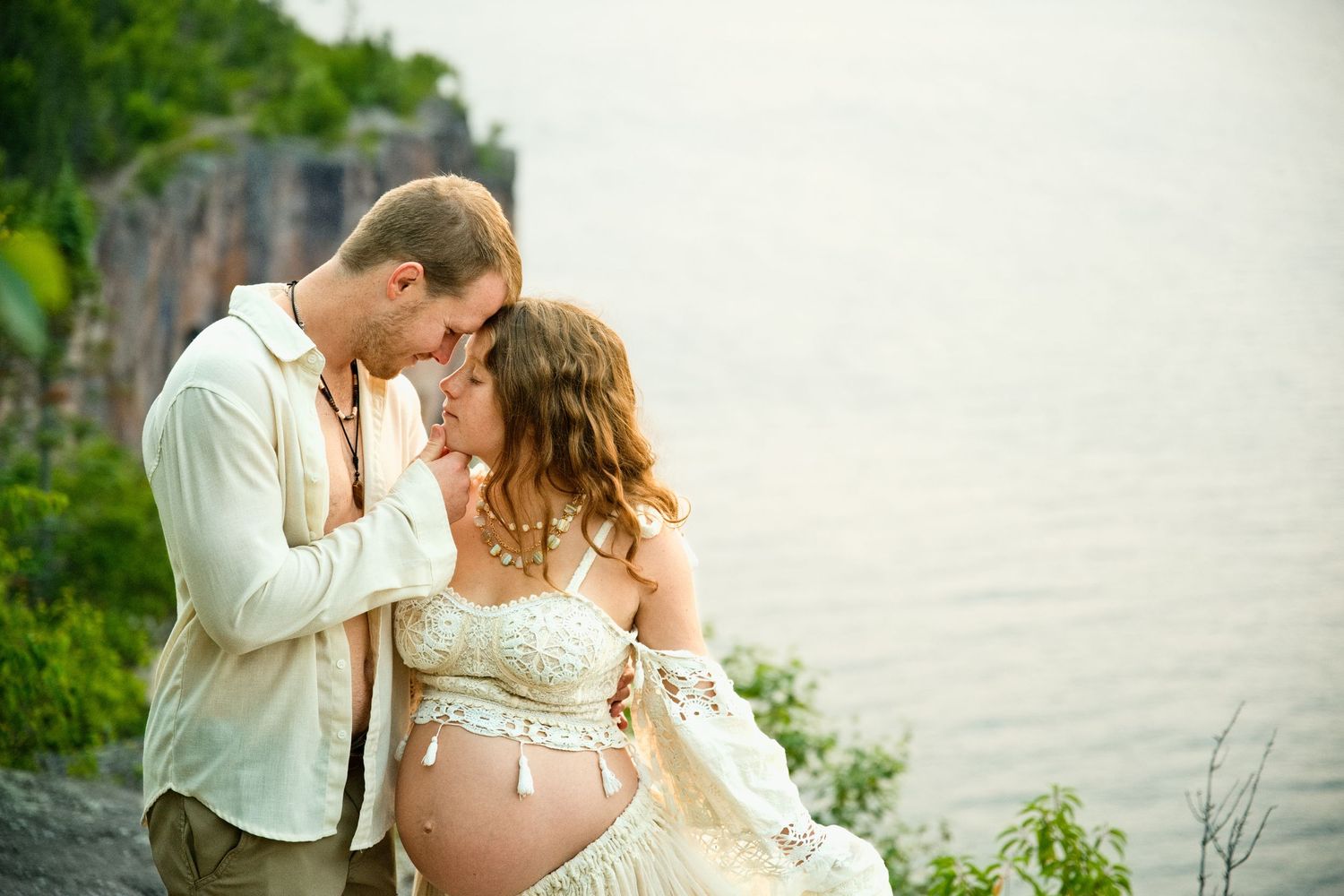 Couple sharing a tender moment outdoors by a lake, woman in lace maternity dress showing baby bump, scenic cliffside backdrop.