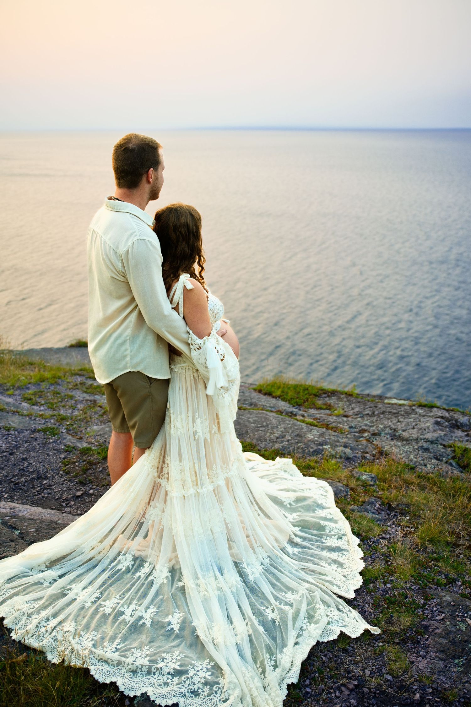 Couple in elegant attire embrace on rocky cliff overlooking calm ocean at golden sunset.