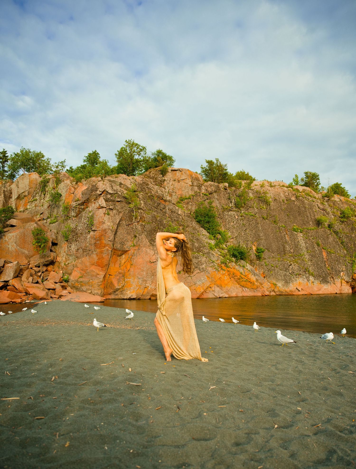 Woman in sheer dress poses on dark sand beach with seagulls, red rock cliffs, and lush green trees backdrop.