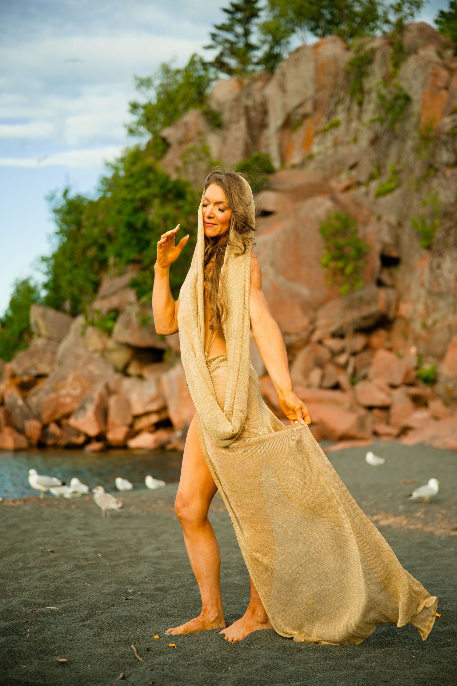 Woman in flowing burlap dress poses barefoot on dark sandy beach with rocky cliffs and seagulls in background.