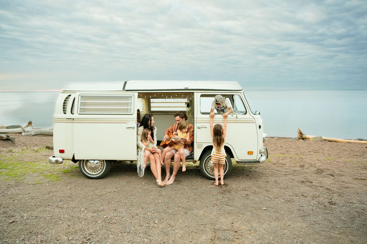 Friends relaxing by a vintage white VW camper van parked on a rocky beach with ocean views on a cloudy day.