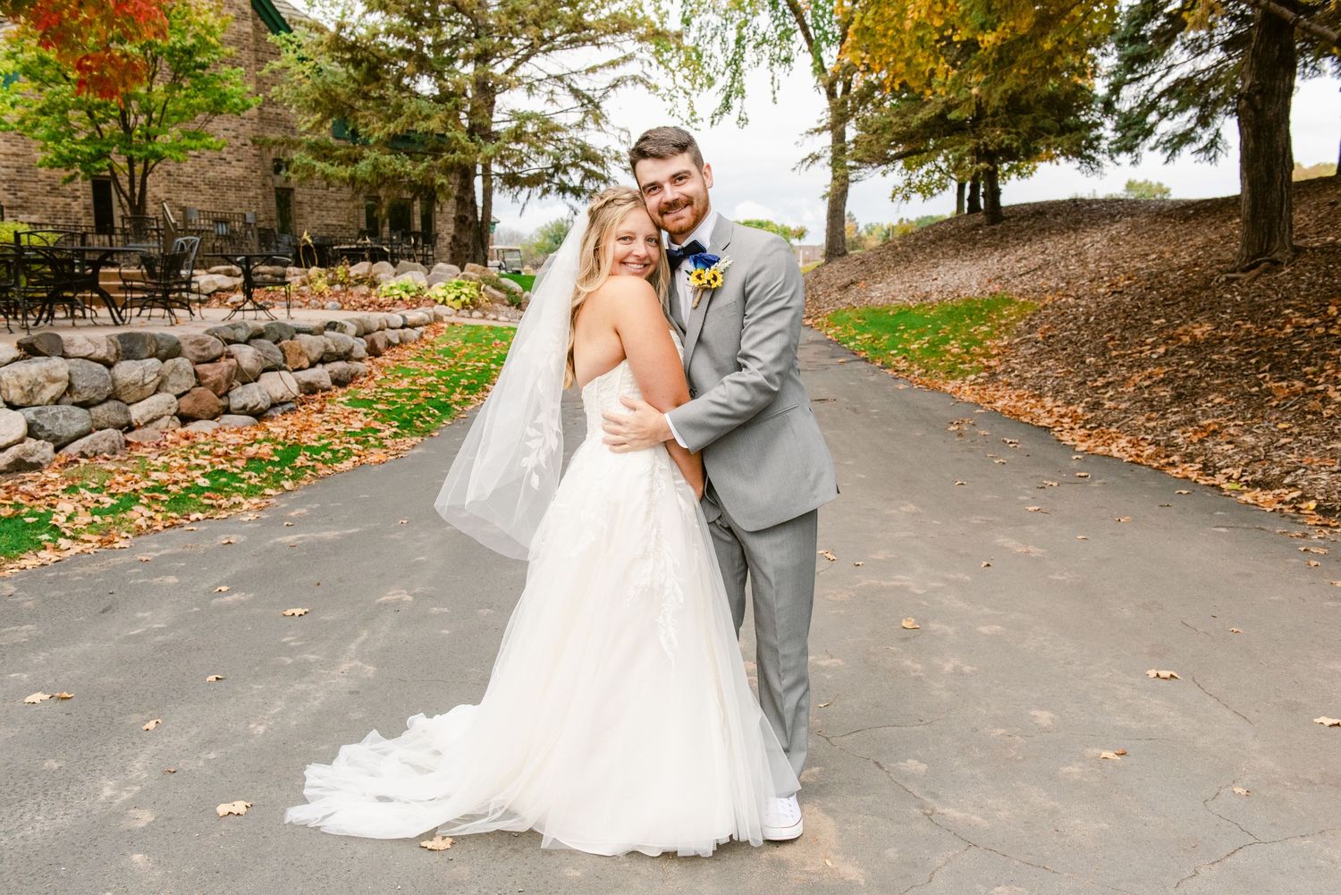 Central MN Wedding Photos | Bride and groom embrace on a scenic path surrounded by vibrant autumn trees and stone walls.