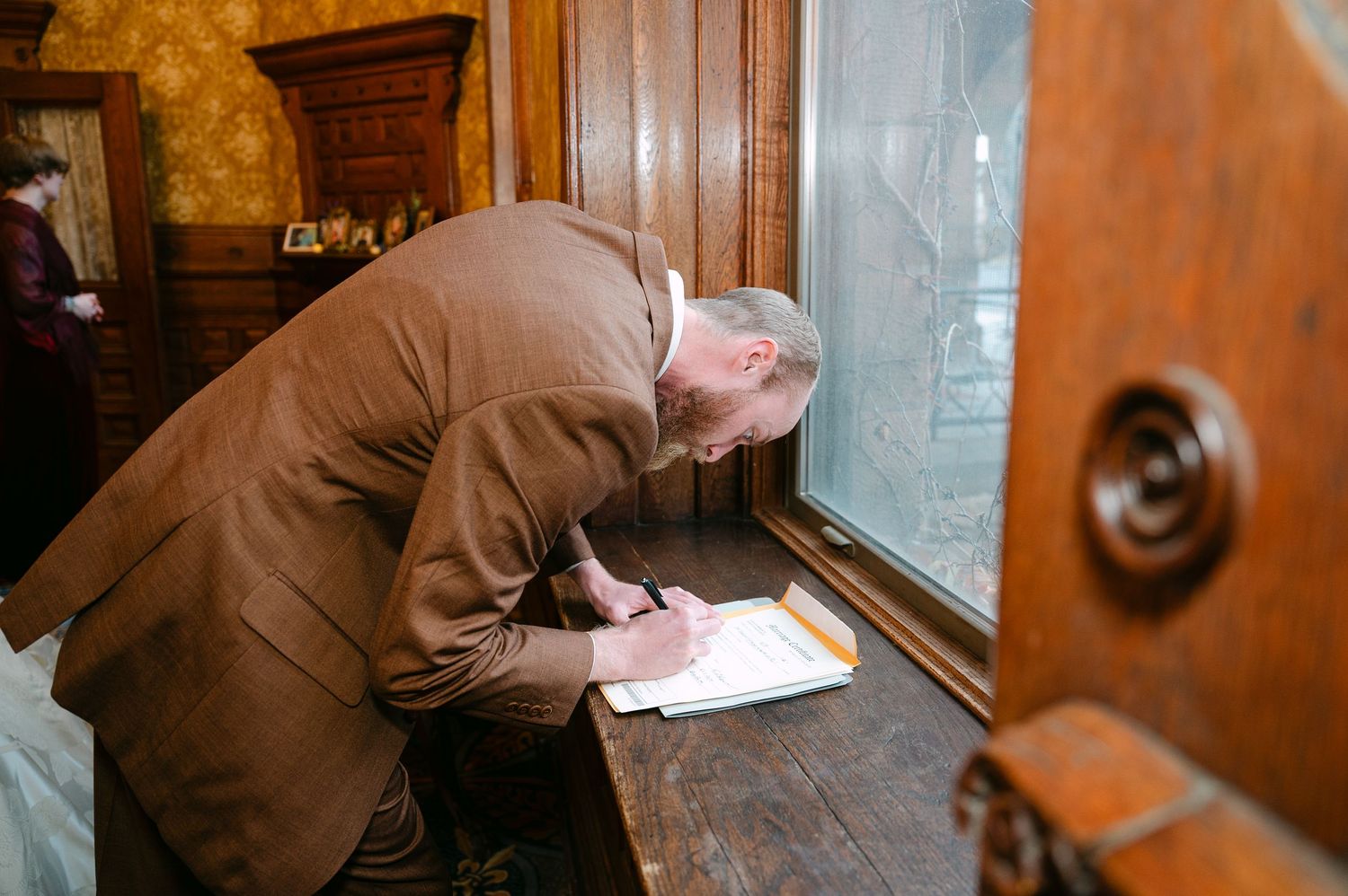 Central MN Wedding Photos | Man in brown suit signing documents at a wooden desk by a window.