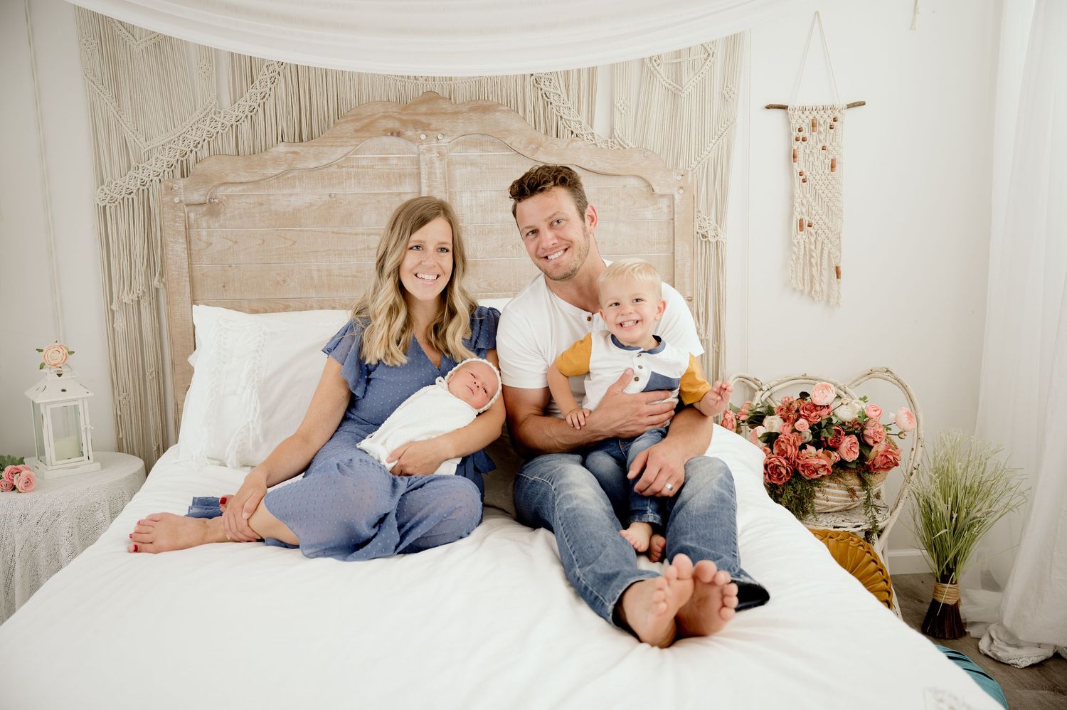 Happy family of four poses on a white bed, parents holding two young children in a bright, bohemian-styled photo studio.