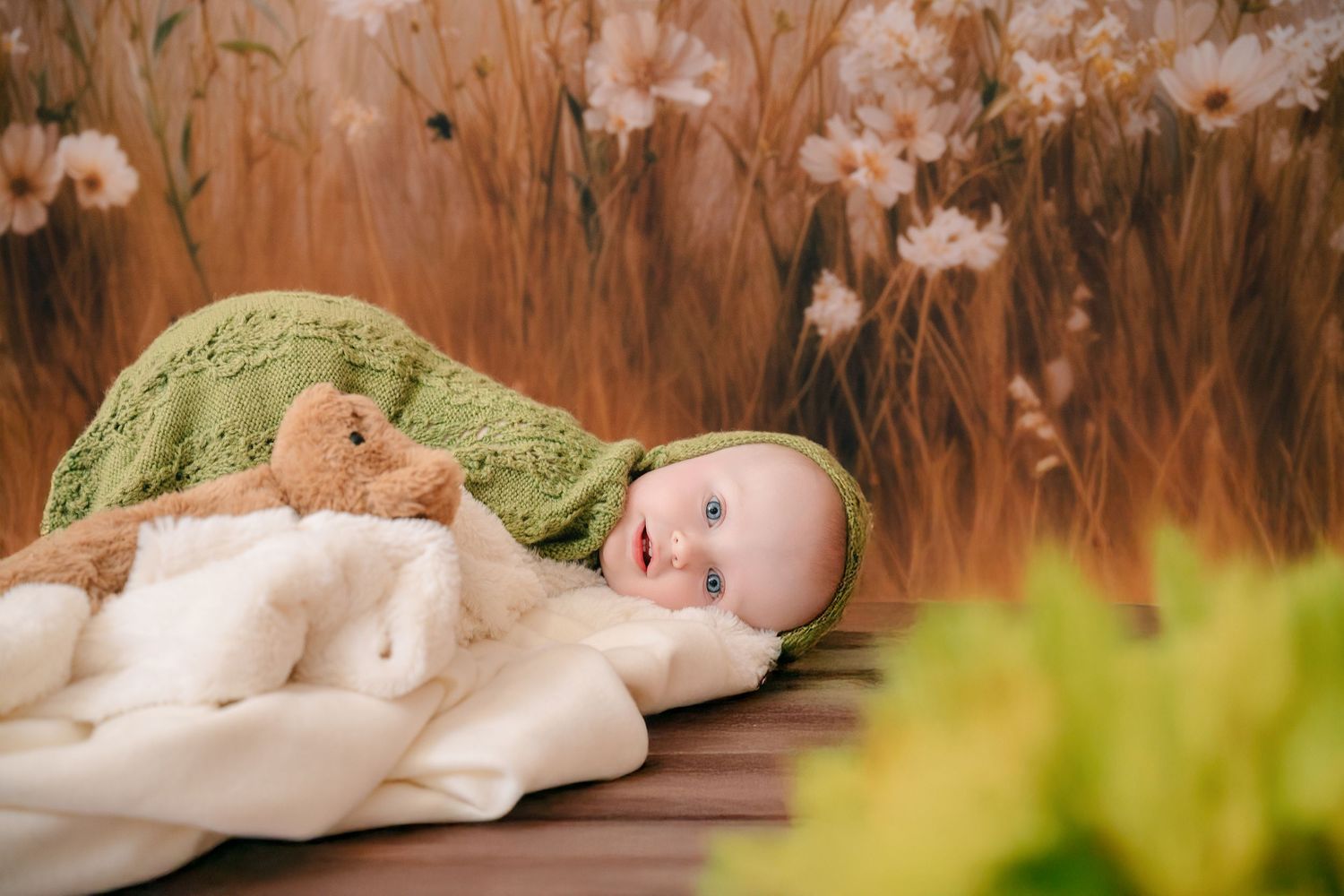 Sweet baby in green knit wrap lying with teddy bear on white blanket against warm floral background. mother day photoshoot