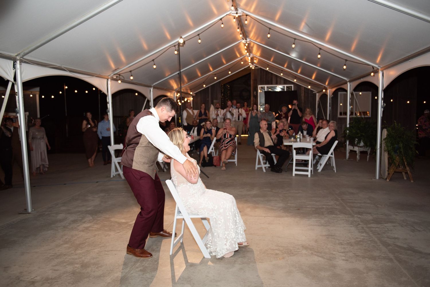 Wide angle shot of a dimly lit wedding tent showing multiple tables and dancing guests under twinkling lights.