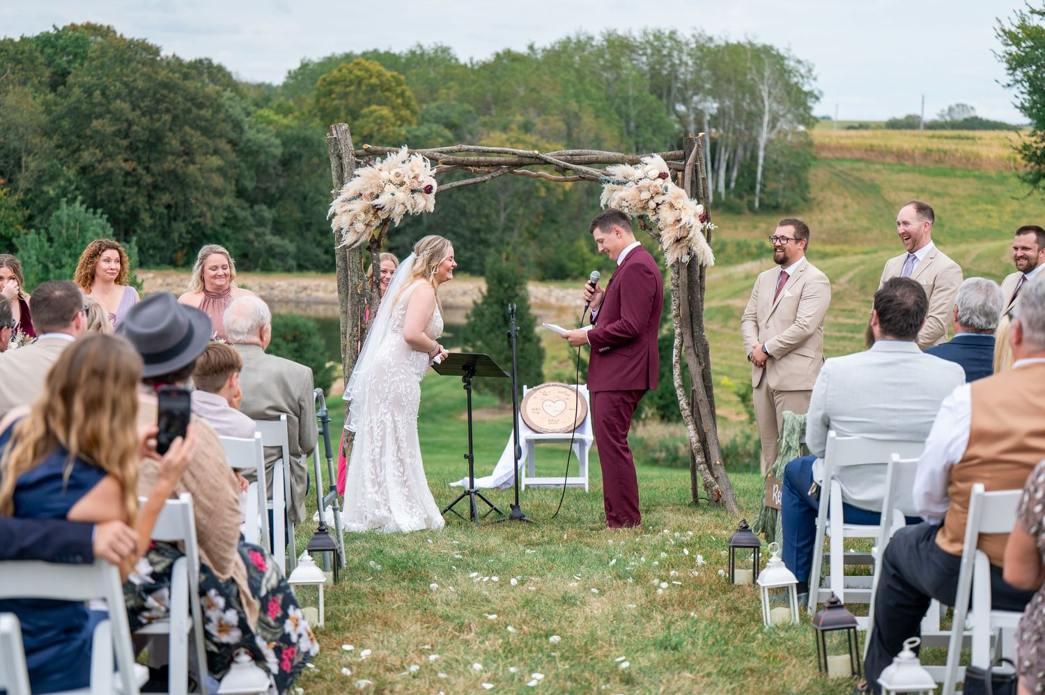 Rustic outdoor wedding ceremony with guests gathered around floral decorated arch and white chairs.