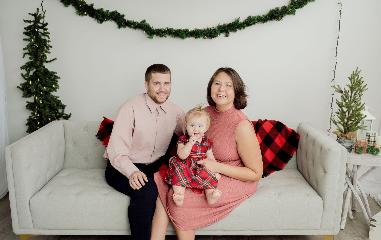 A family poses on a gray couch decorated with festive holiday greenery and buffalo plaid accents.