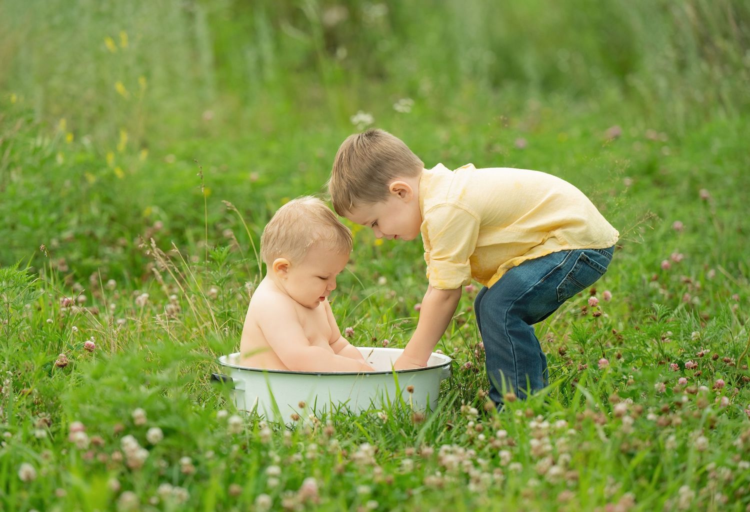 A curious child in blue jeans and yellow shirt crawls through green grass while exploring outdoors on a sunny day. Family Portraits & Milest