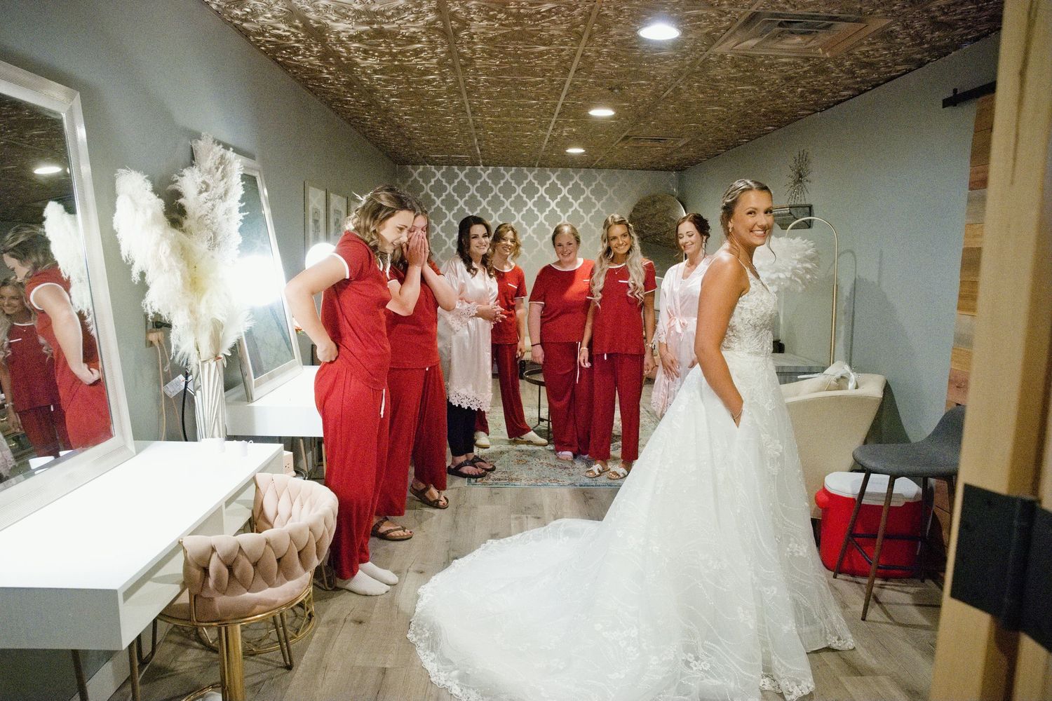Bridesmaids in red outfits react emotionally as bride in a lace wedding gown reveals her look in a bridal suite.