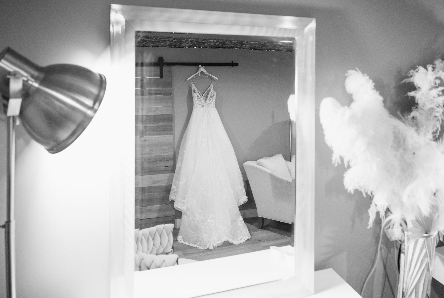 Black and white photo of elegant lace wedding dress hanging on a barn door reflected in a mirror.