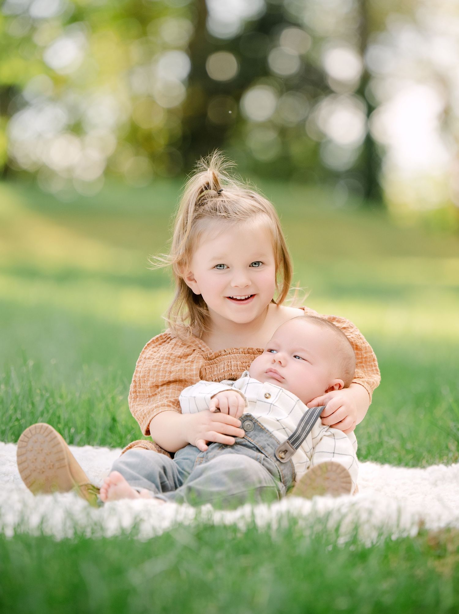 A smiling toddler girl holds a newborn baby sibling on a blanket in a sunny green park.. Minnesota milestone photographer