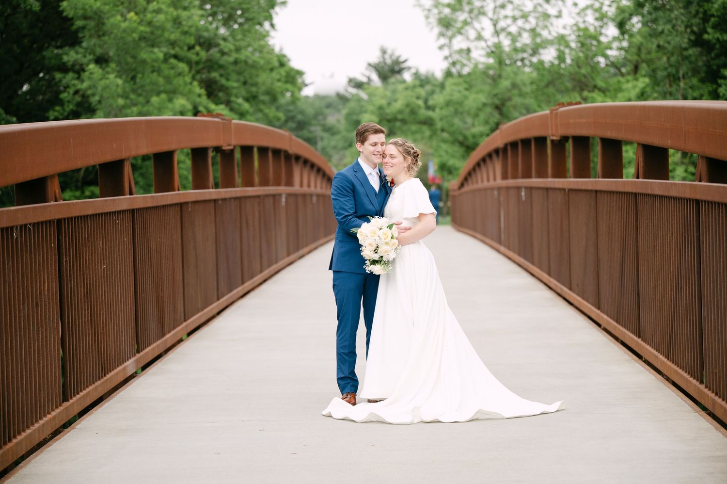 Romantic wedding couple embraces on a wooden bridge surrounded by lush greenery and trees.