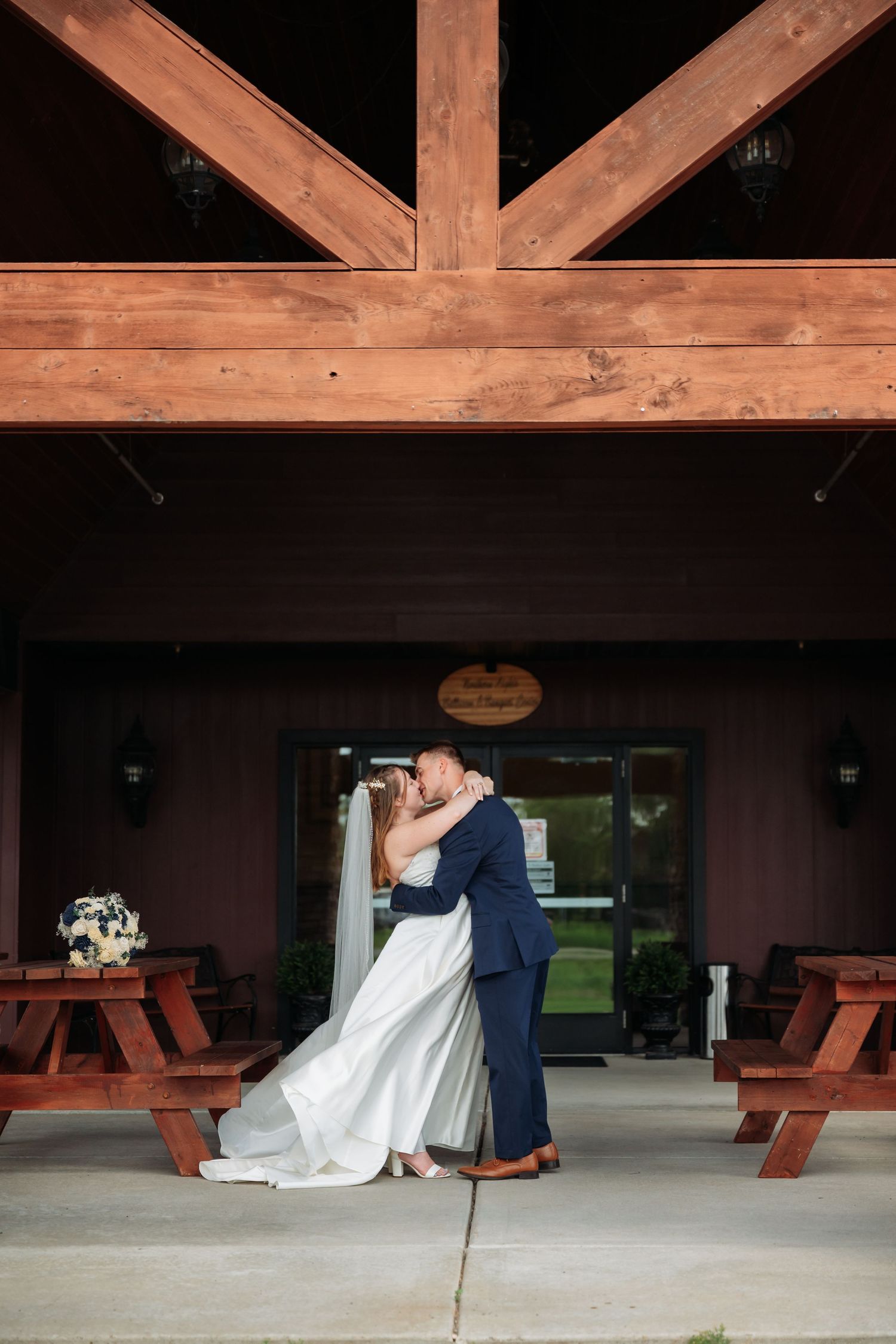 Wedding couple embracing under rustic wooden beams with picnic tables in background. Pease Minnesota Wedding at Northern Lights Ballroom & B