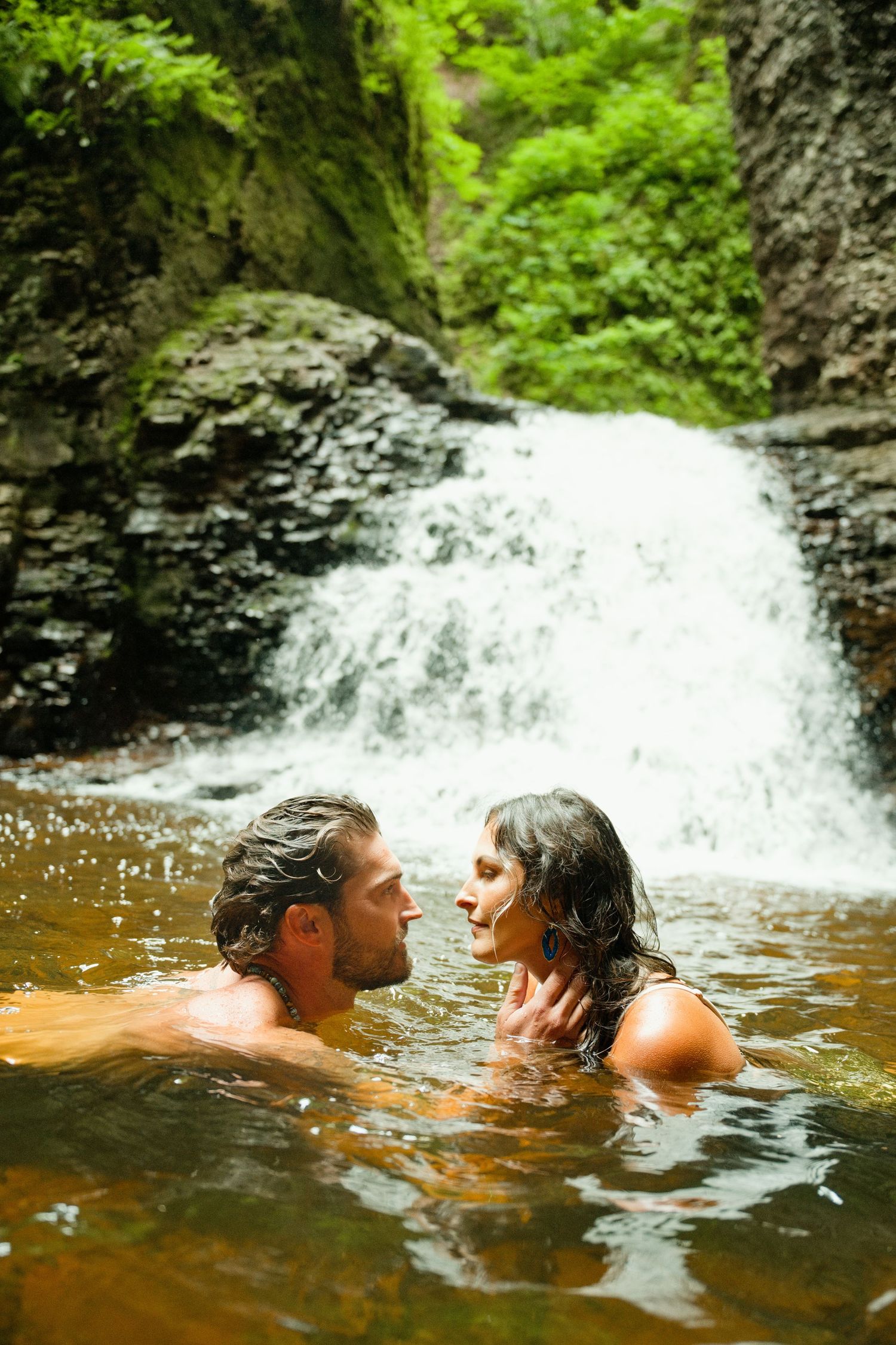 Romantic couple embracing in a natural pool with a lush waterfall cascading over mossy rocks in the background.