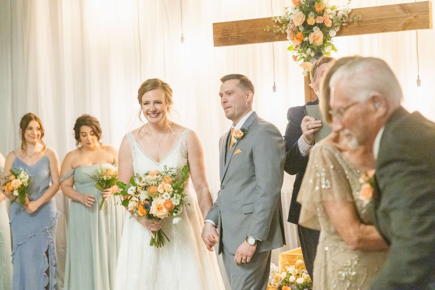 Bride and groom smile with bridesmaids holding peach and orange bouquets after ceremony.