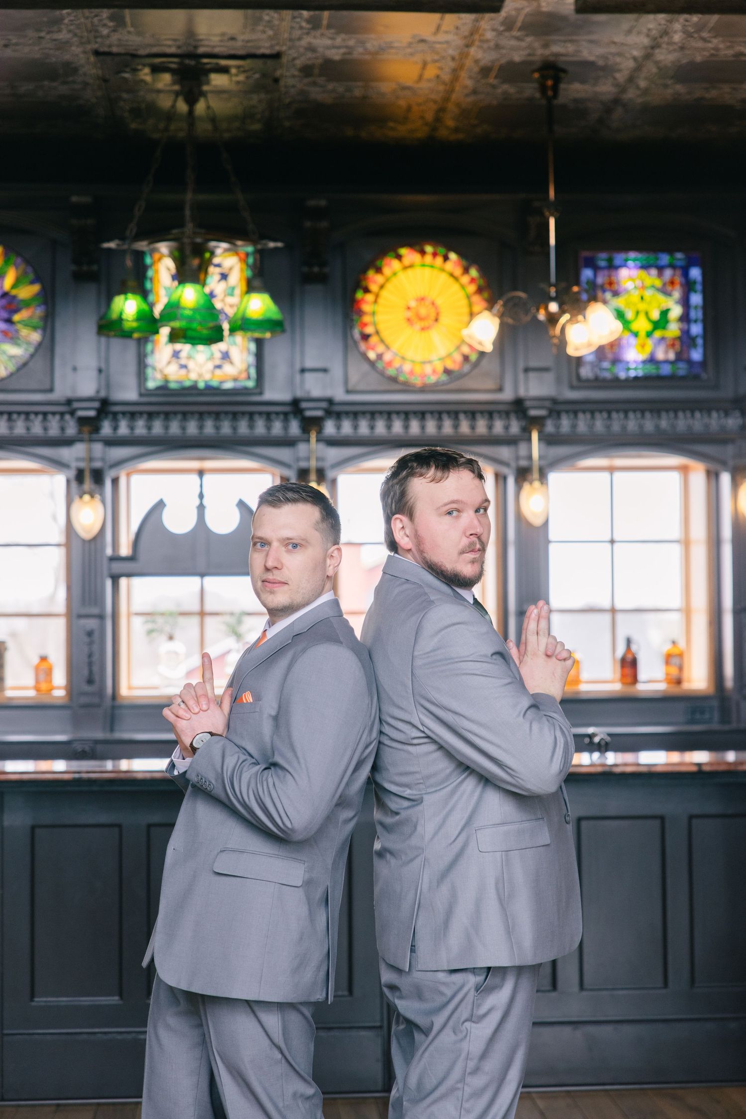 Two groomsmen in matching gray suits pose back-to-back in a vintage bar with stained glass windows.