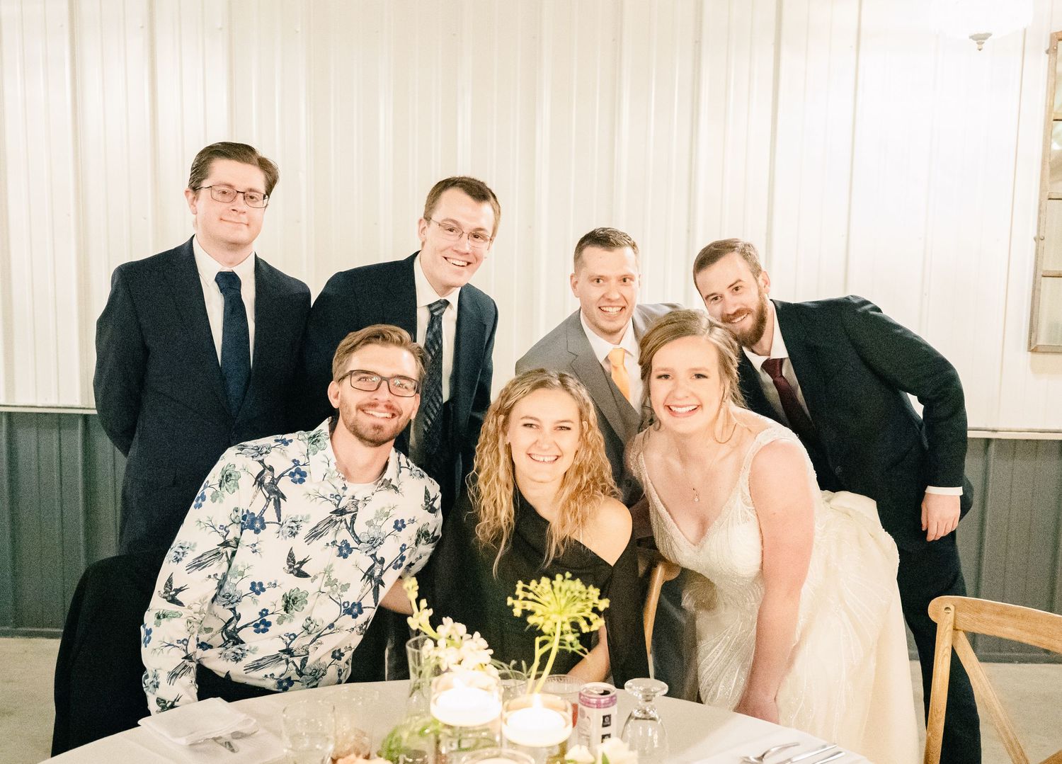 Bride and groom pose with a group of smiling friends at a wedding reception table.