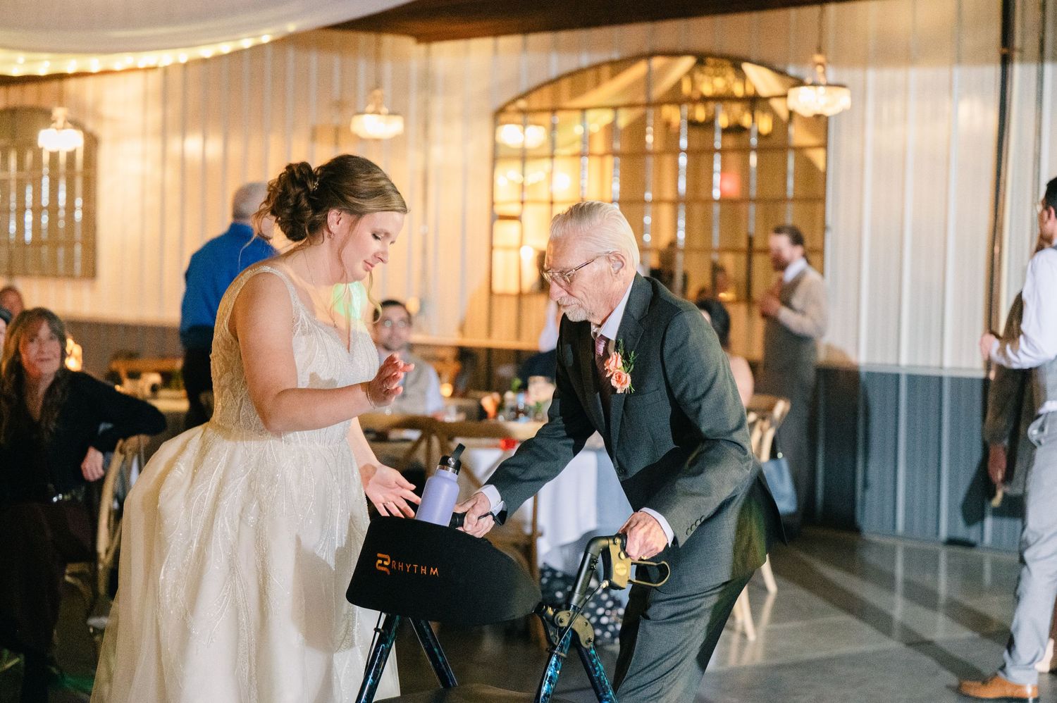 Bride in white gown interacting with elderly guest using walker at wedding reception.