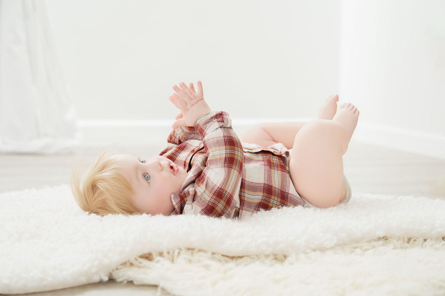 Central MN Milestone Photography - Smiling baby in plaid shirt lying on white fluffy rug with legs raised.