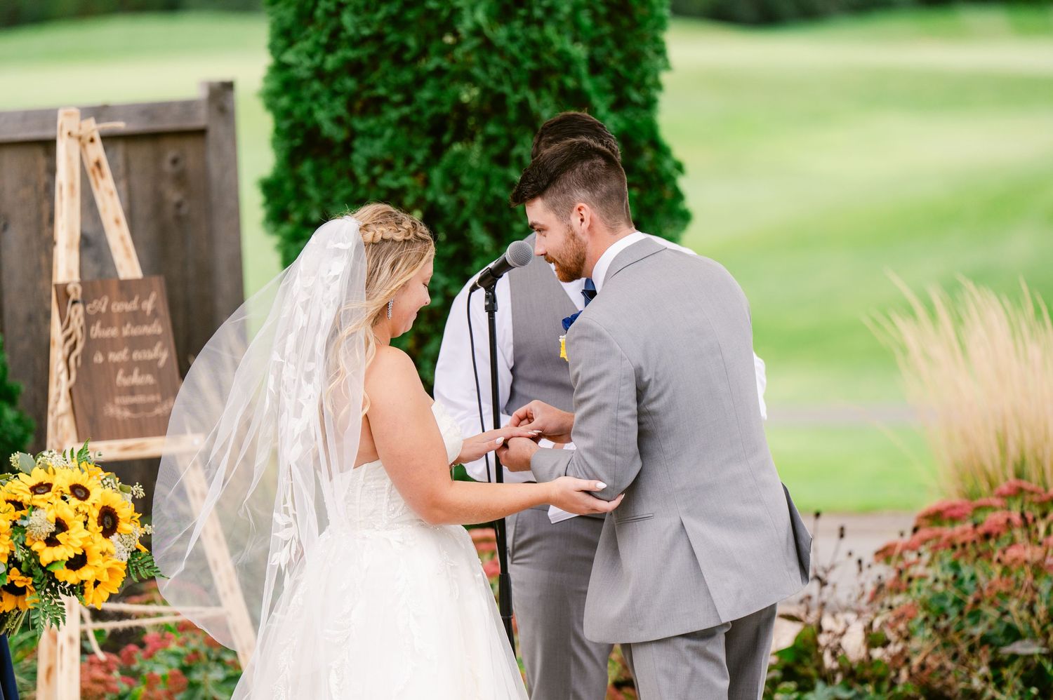 Central MN Wedding Photos - Bride and groom exchange rings during outdoor wedding ceremony surrounded by sunflowers and greenery.
