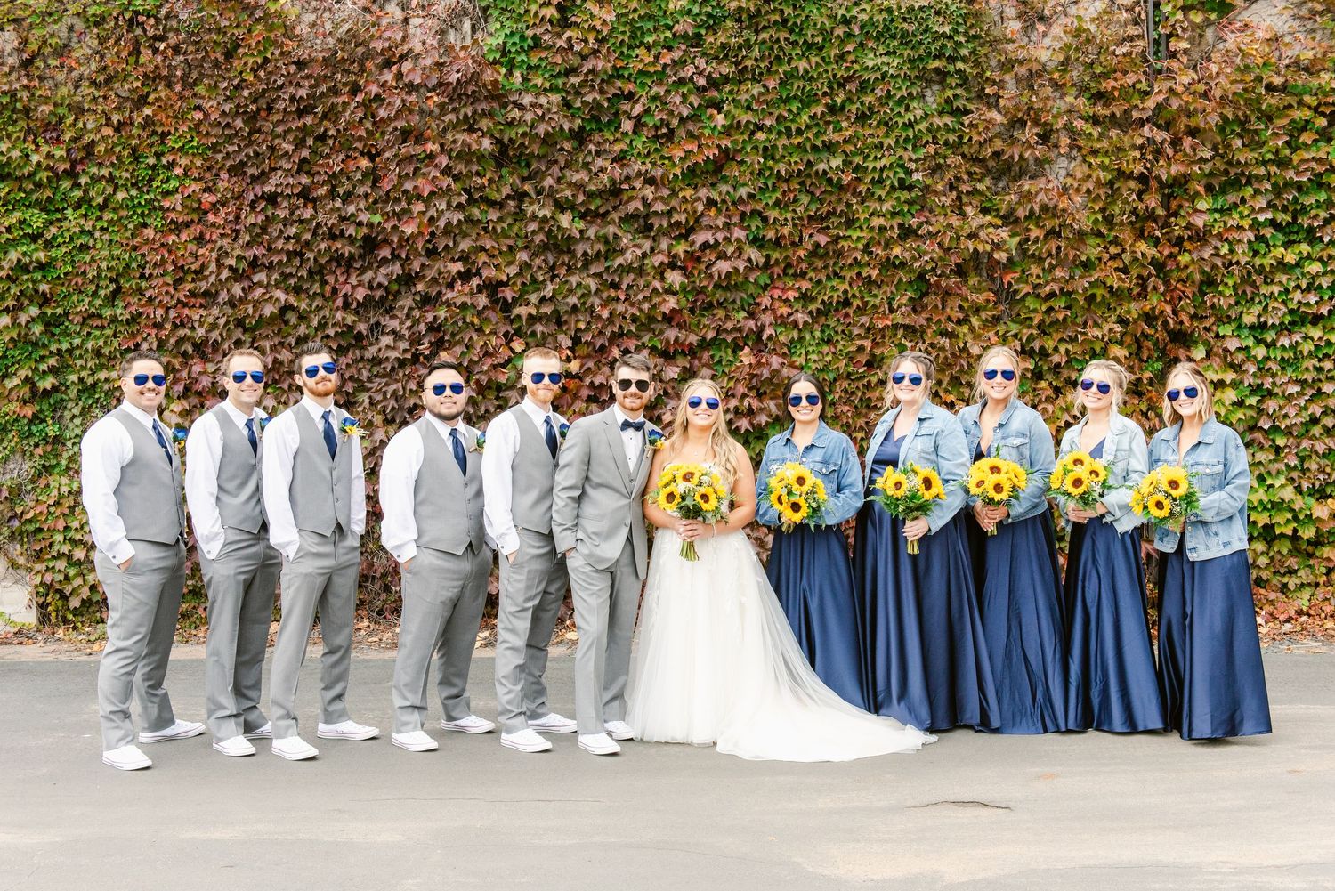 Central MN Wedding Photos | Bridal party wearing fun sunglasses poses with sunflower bouquets against colorful ivy backdrop.