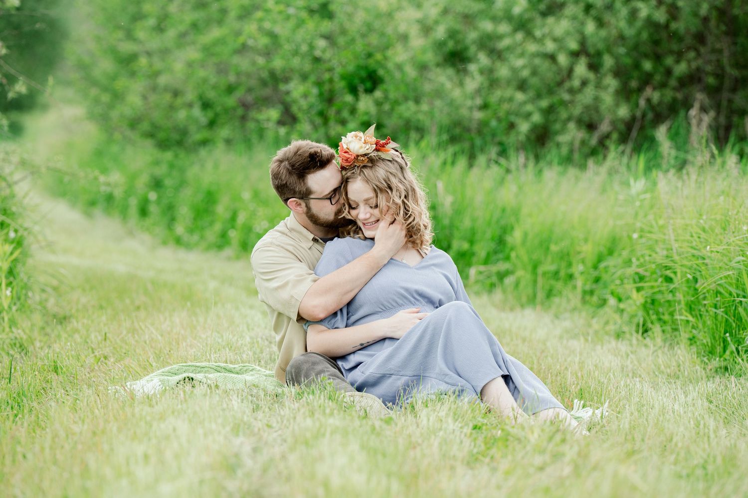A couple sits embracing in a sunlit meadow with tall green grass and natural surroundings.