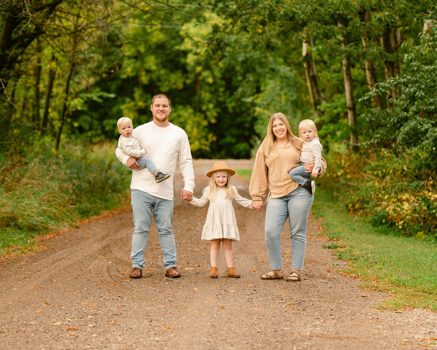 A family walks together on a tree-lined dirt path through lush green woods on a sunny day.