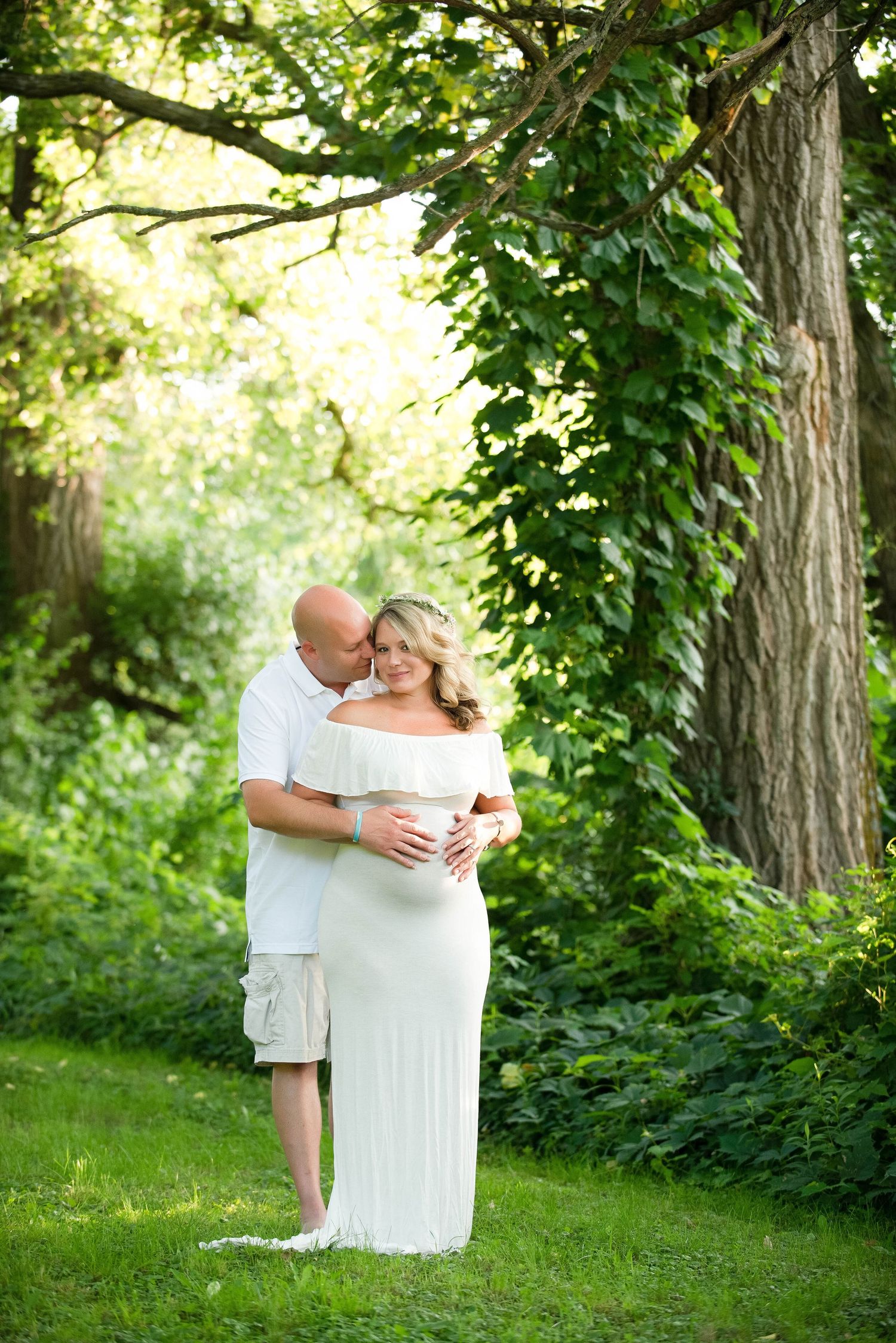 A couple shares an intimate embrace on a grassy lawn surrounded by lush summer trees and greenery.