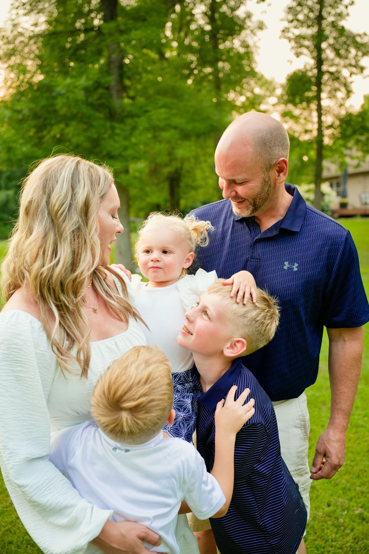 A happy family of five with a blonde mother, bald father, and three young children sharing a warm moment outdoors.