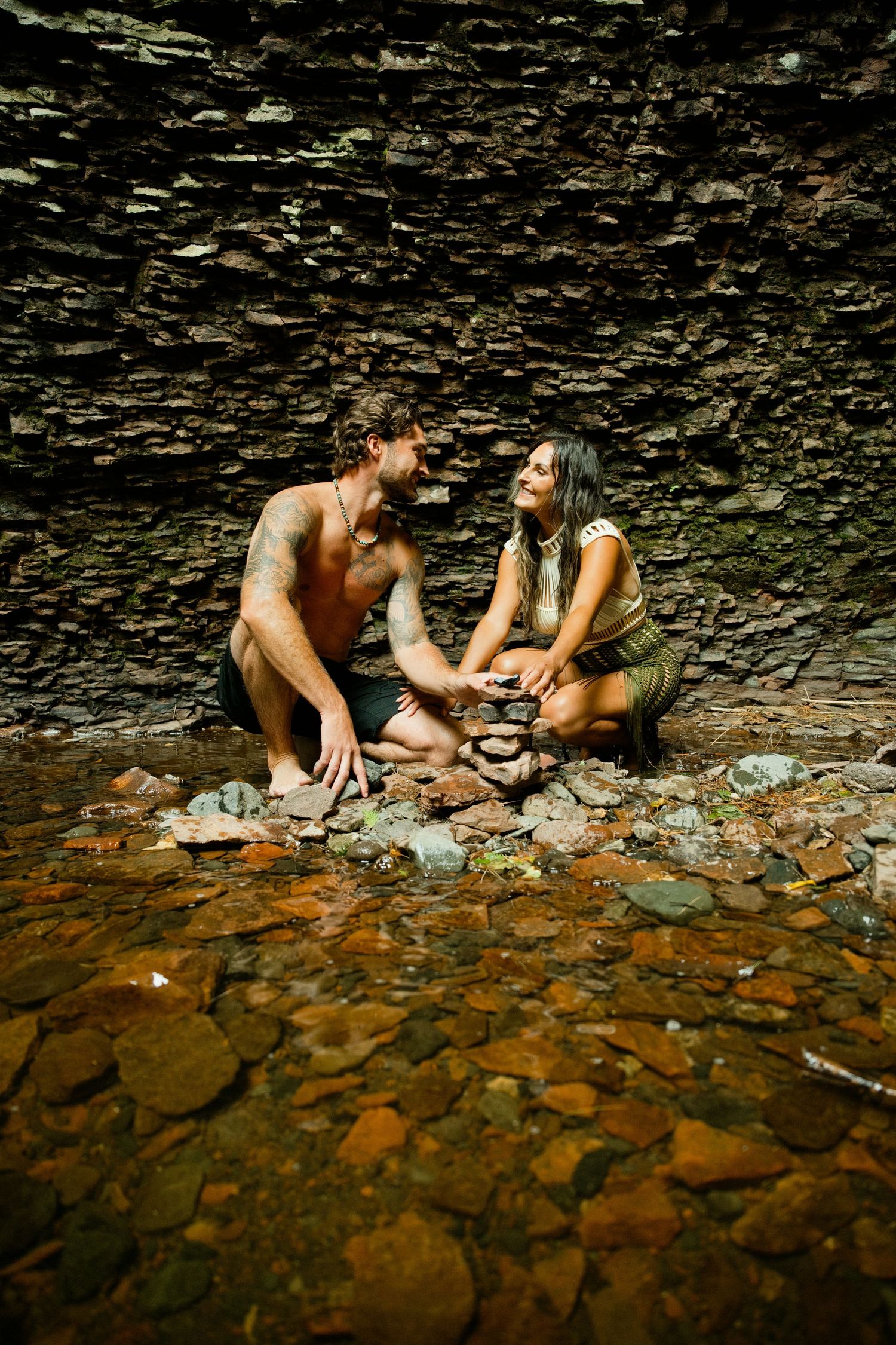 Couple stacking rocks together beside shallow stream against textured stone wall backdrop.