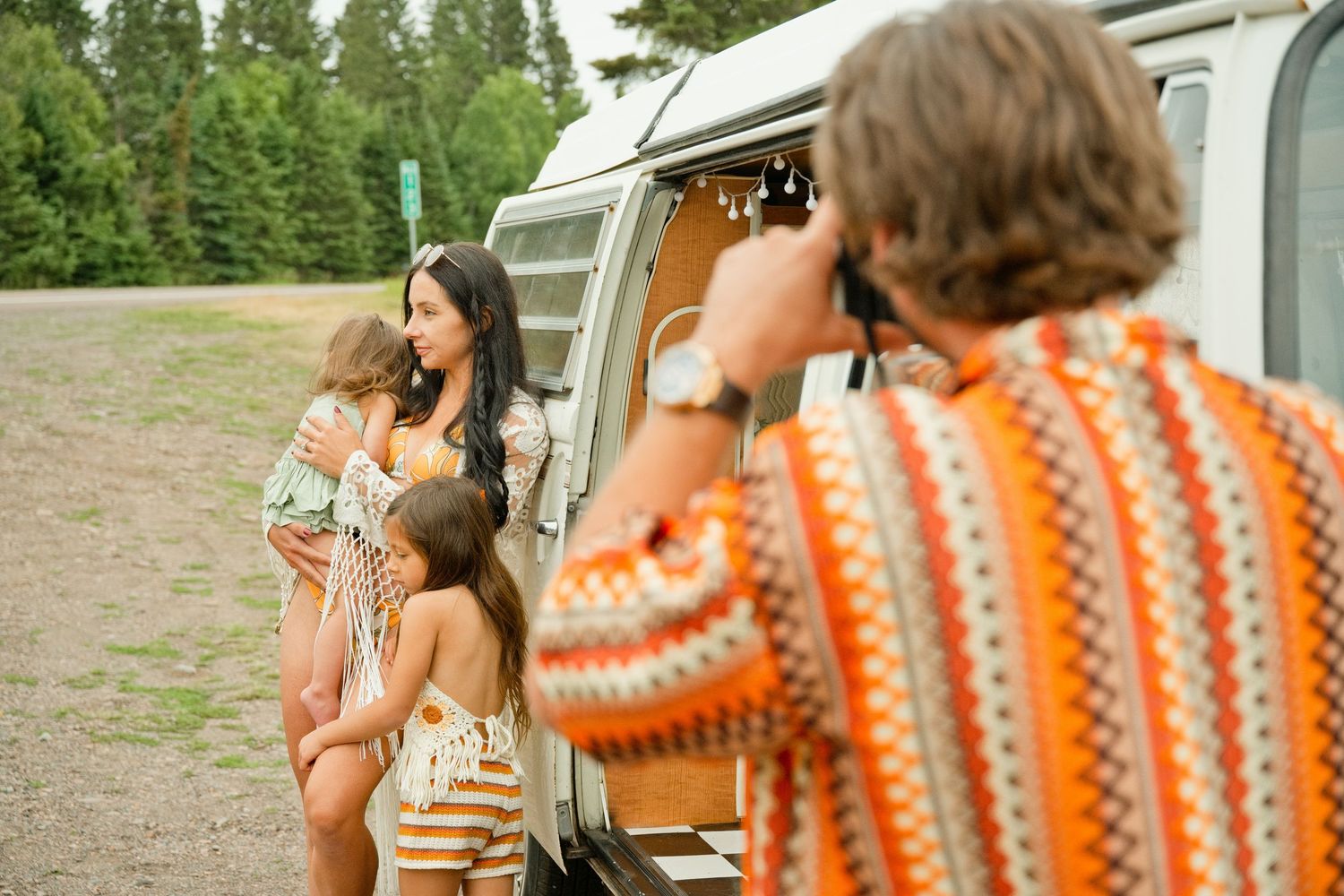 Man photographing two women and a child posing by a vintage van at a forest campsite.