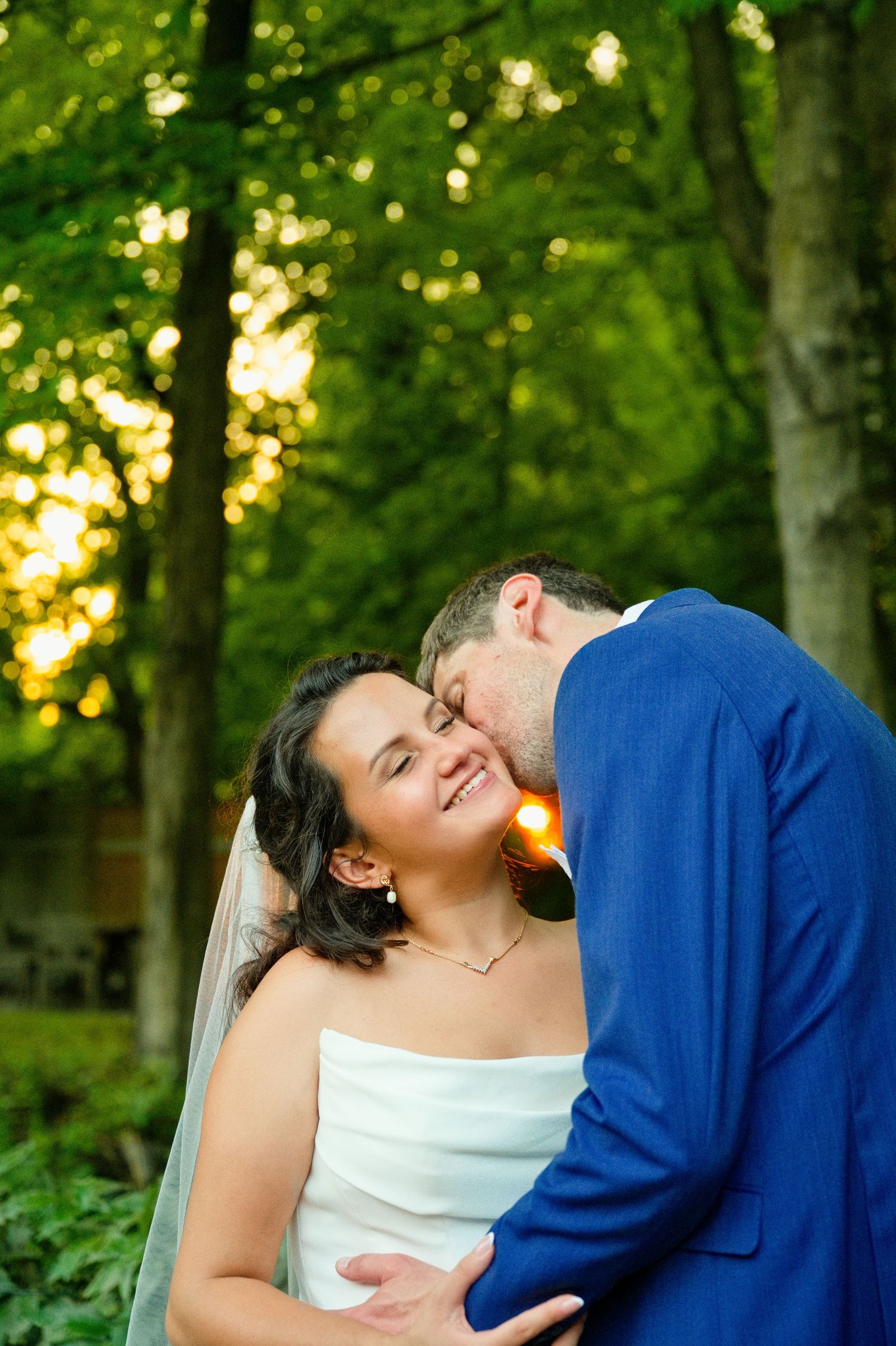 Groom in blue suit kissing bride in white dress and veil, smiling outdoors in lush green forest setting.