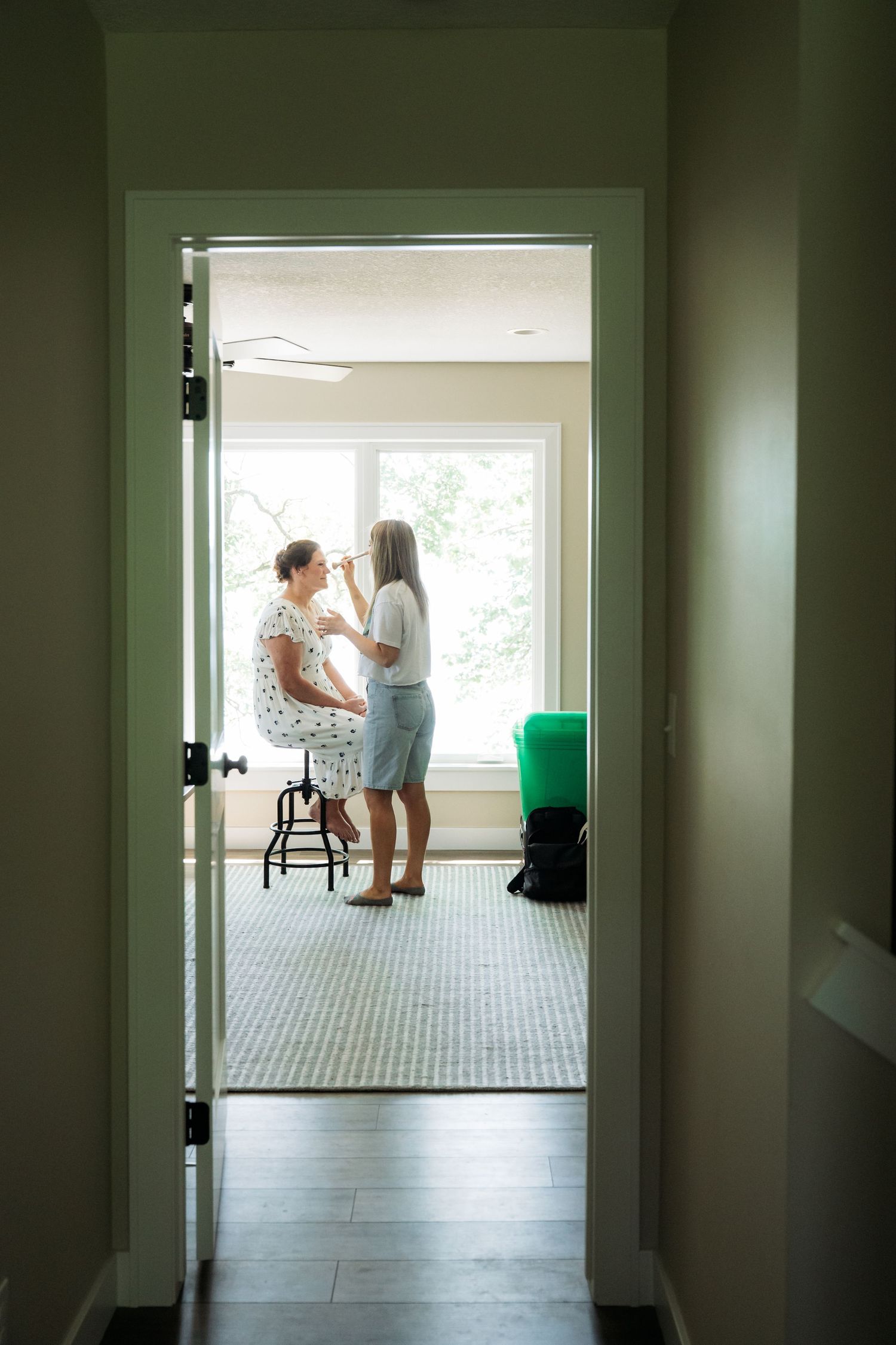 Two women getting ready indoors, one applying makeup to the other near a bright window in a modern room.