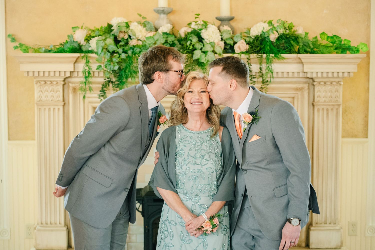 Two grooms in gray suits kiss a woman in a teal dress on the cheek near a floral fireplace.